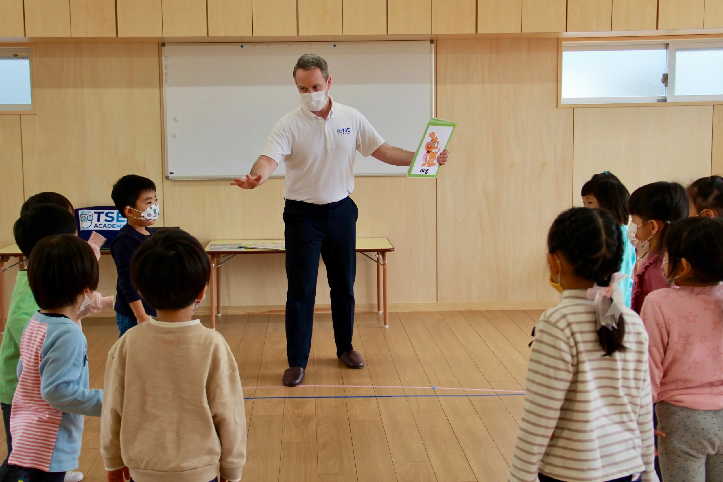 教室で教師が子供たちにカードを見せている。子供たちはマスクを着用し、一列に並んでいる。