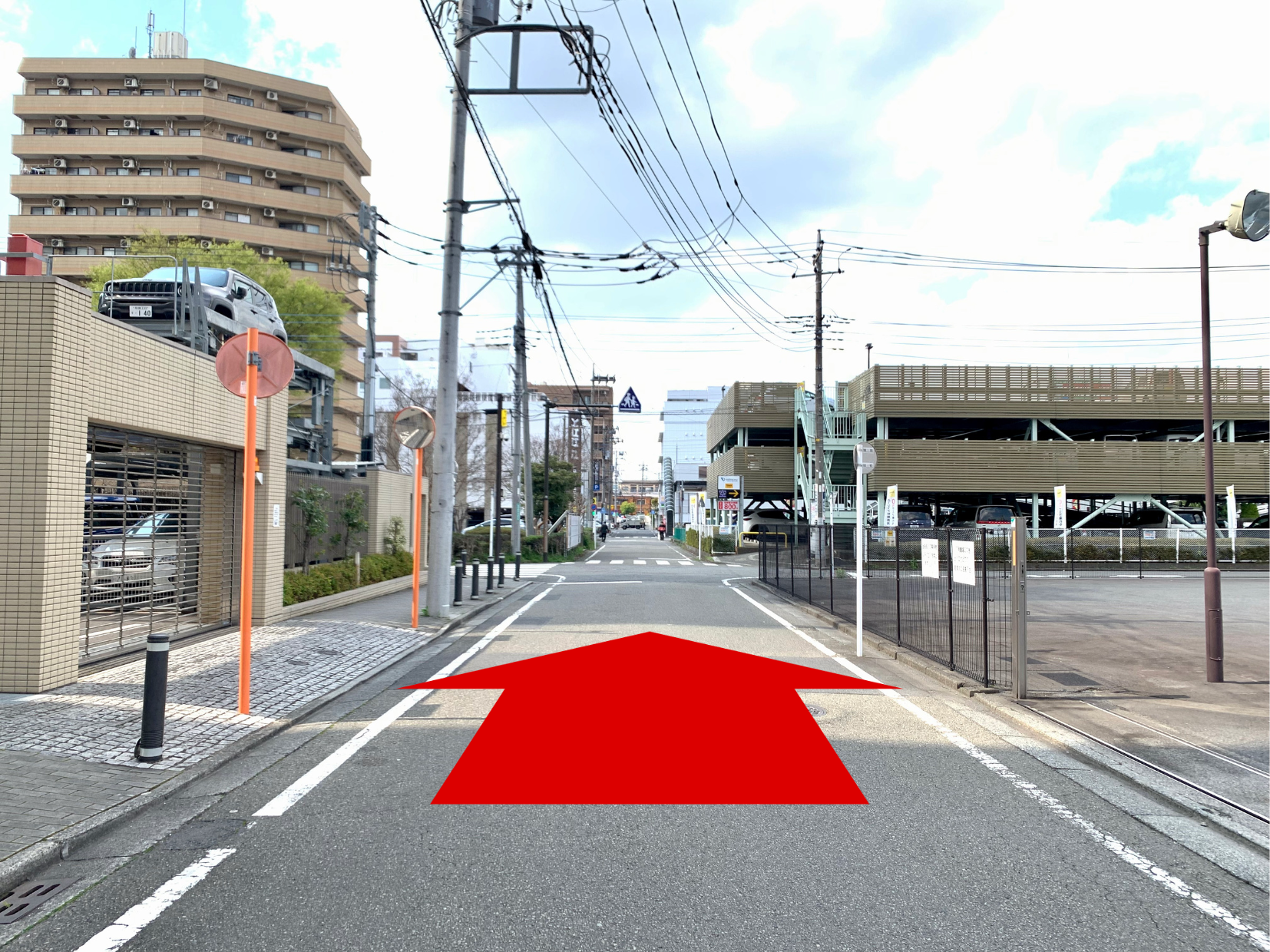 赤い矢印が示す道路の光景。左側にビル、右側に駐車場があり、電信柱や電線が見える。晴れた青空が広がっている。