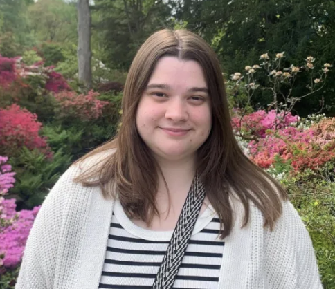 A young woman with long brown hair smiling, standing outdoors near water with rocks and houses in the background, wearing a colorful fleece jacket.