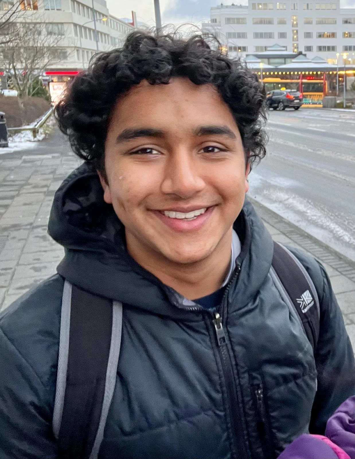 Close-up of a young man with curly black hair smiling outdoors in winter, wearing a black puffer jacket and a backpack, with snow and a city street in the background.