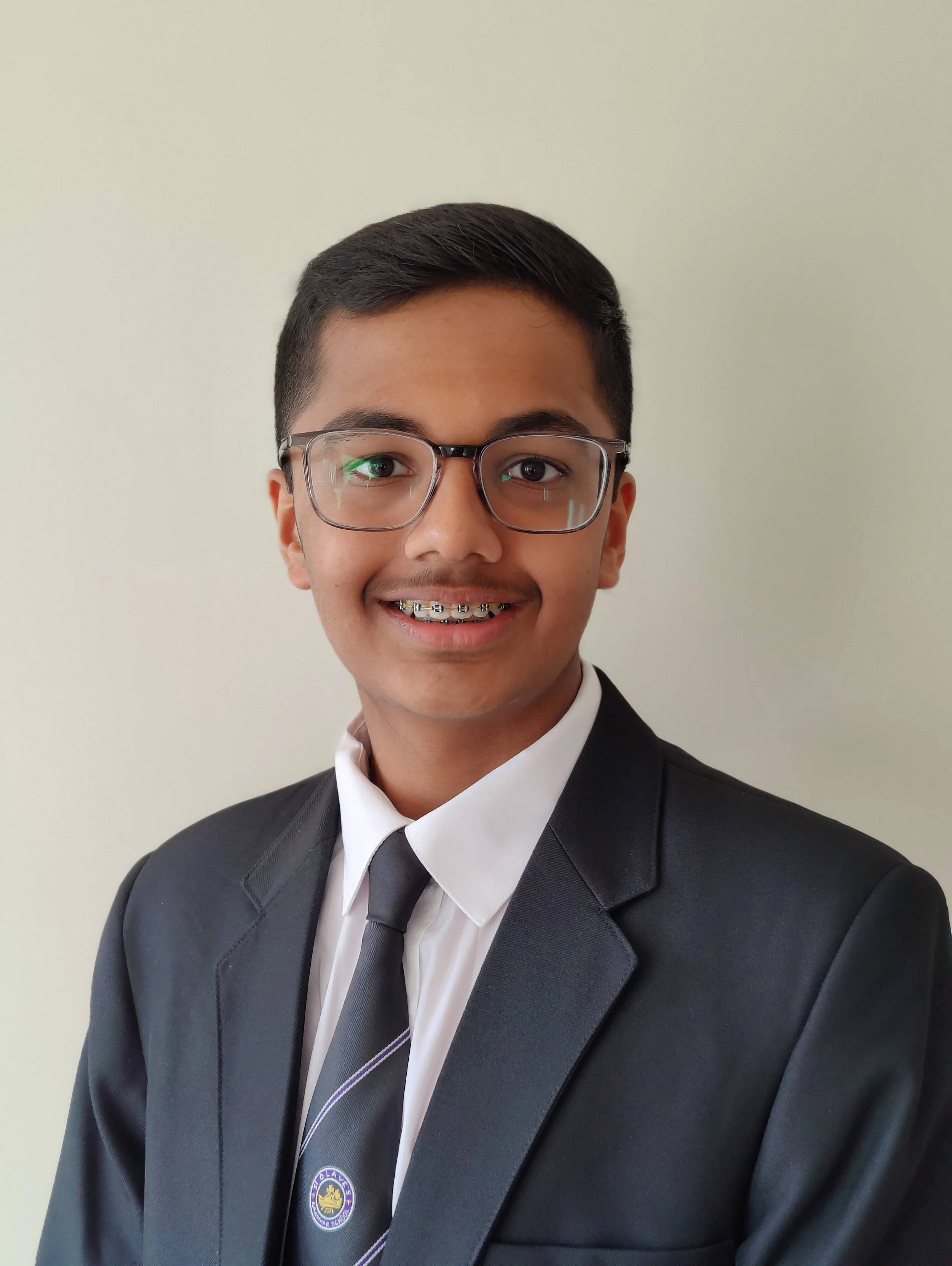 A young man in a black suit, white shirt, and striped tie, smiling, wearing glasses, against a plain light background.