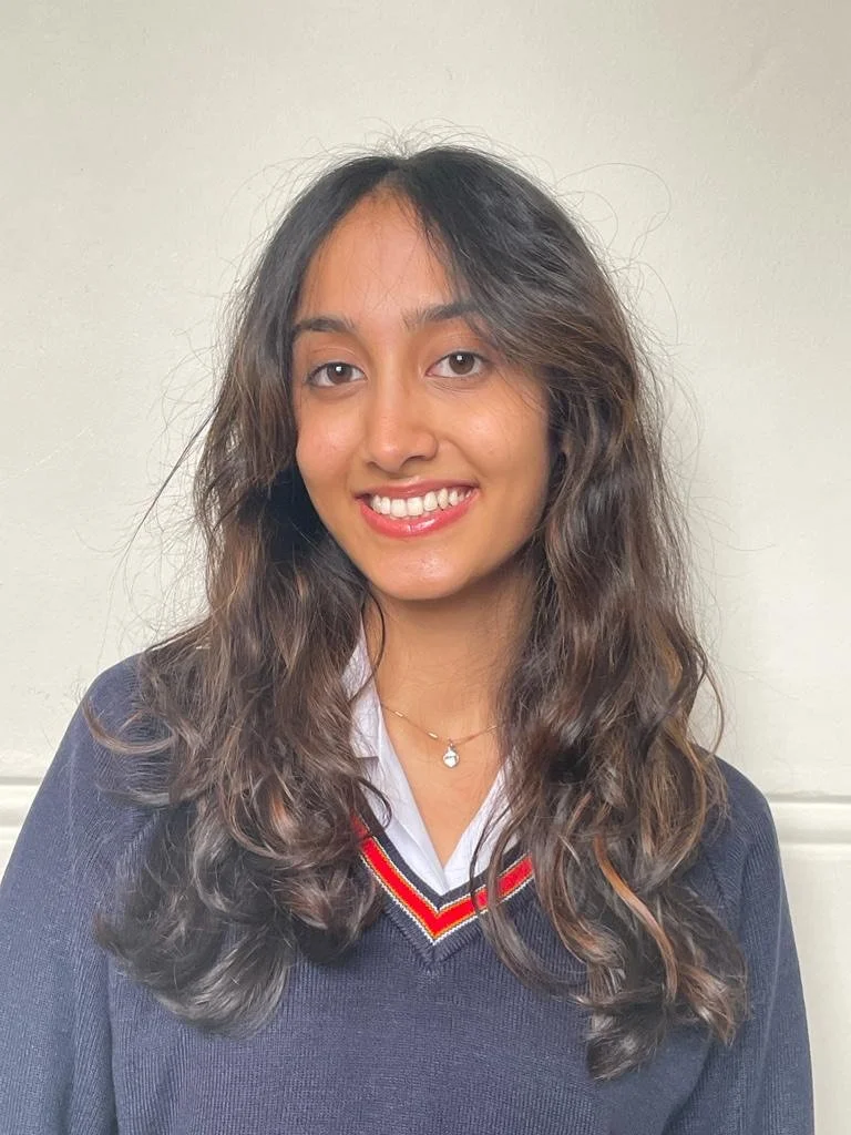 A young woman with long, wavy brown hair smiling, wearing a navy school uniform sweater with red, white, and yellow stripes, and a white collared shirt underneath, standing against a light-colored wall.
