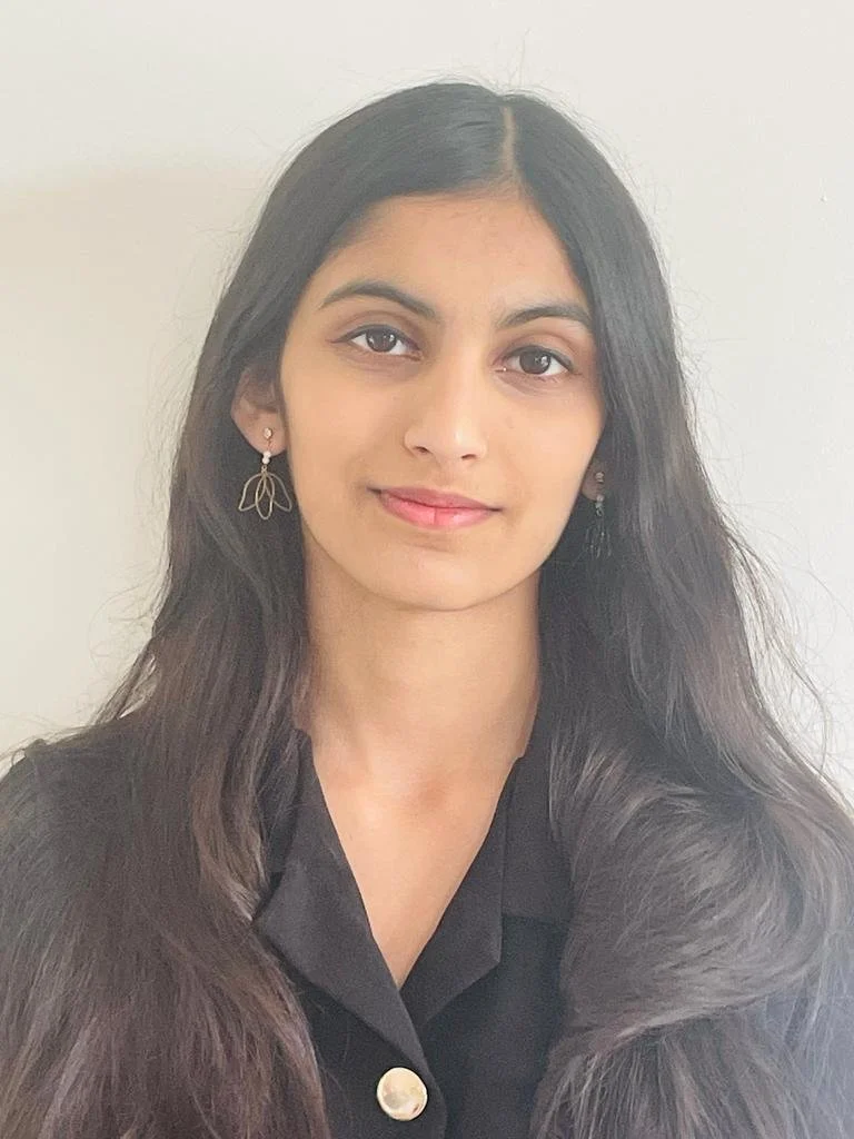 A young woman with long dark hair, wearing earrings, a black top, and smiling softly in front of a plain white background.