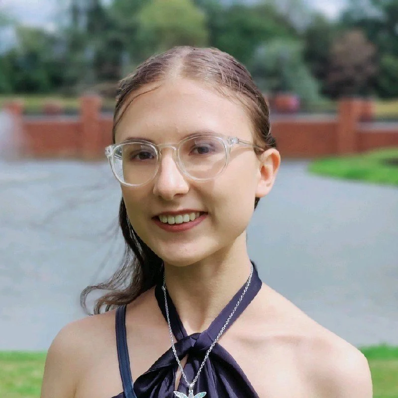 Young woman with clear glasses and long brown hair outdoors near a pond, smiling at the camera.