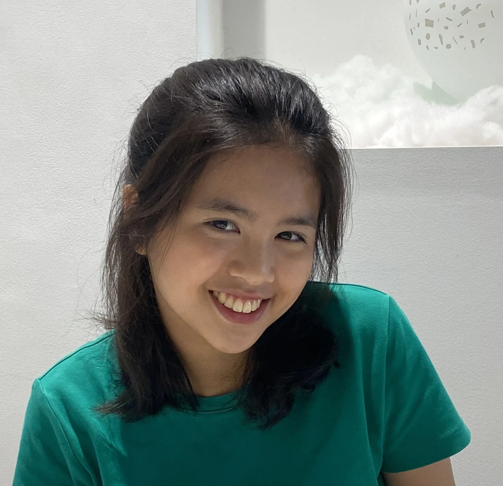 A young woman with dark hair smiling and wearing a green shirt, sitting indoors near a window with a cloudy sky visible outside.