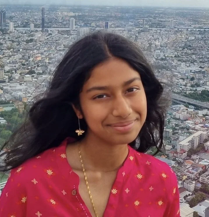 Young woman with long black hair smiling, wearing a red traditional dress with yellow floral embroidery, earrings, and a gold necklace, standing on a high vantage point overlooking a city skyline.