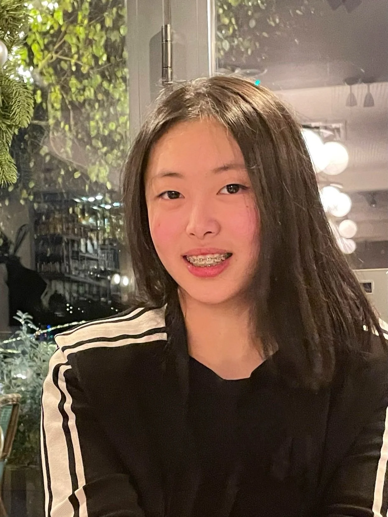 Young woman with shoulder-length dark hair and braces smiling at the camera, wearing a black jacket with white stripes on the sleeves, indoors at a restaurant or cafe with dim lighting and decorative ceiling lights.