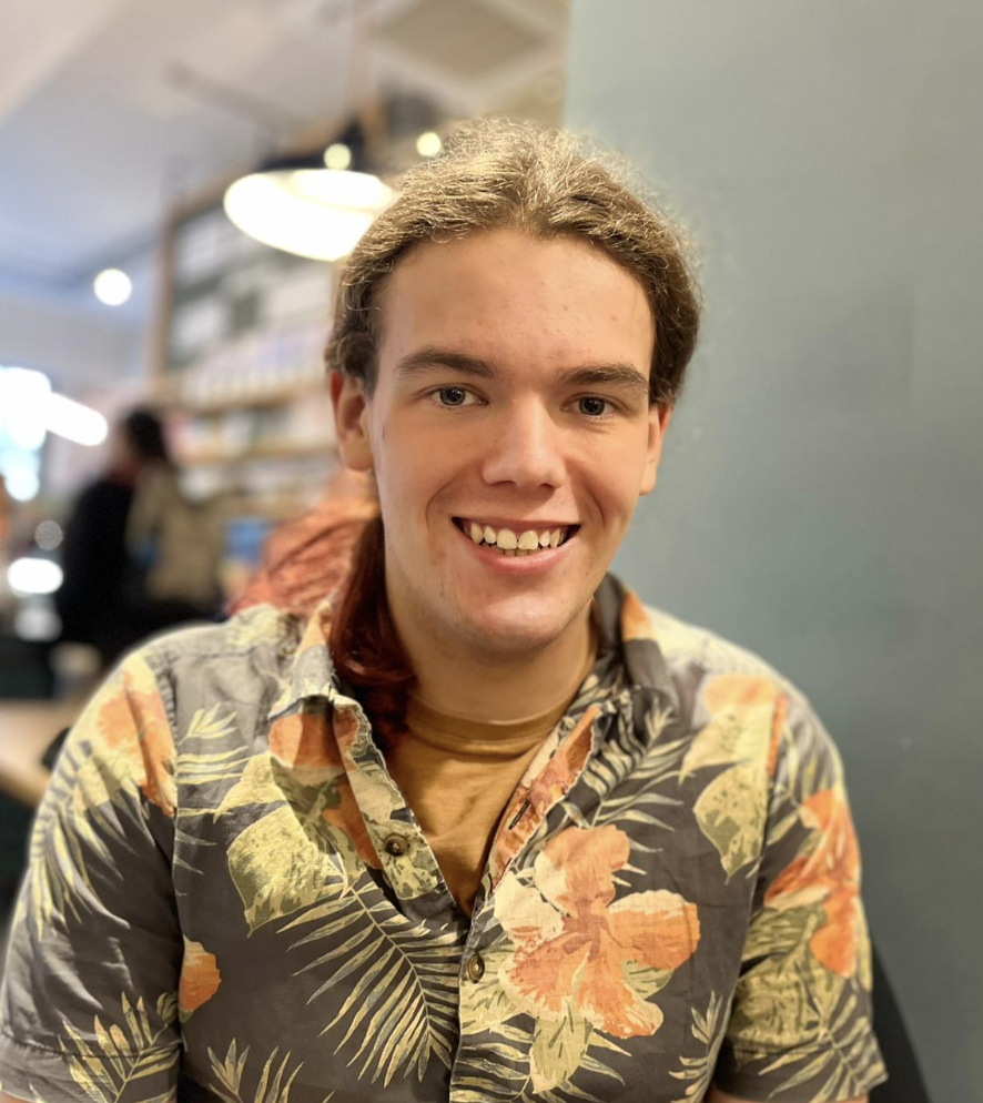 Young person with long hair that is brown at the top and red at the tips, smiling at the camera indoors with shelves of comic books or graphic novels in the background.