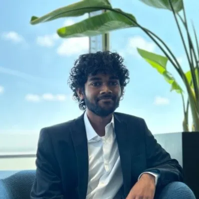 A man with curly hair and a beard wearing a dark suit jacket and white shirt, sitting indoors near a large plant with broad leaves and a window showing a blue sky with clouds.