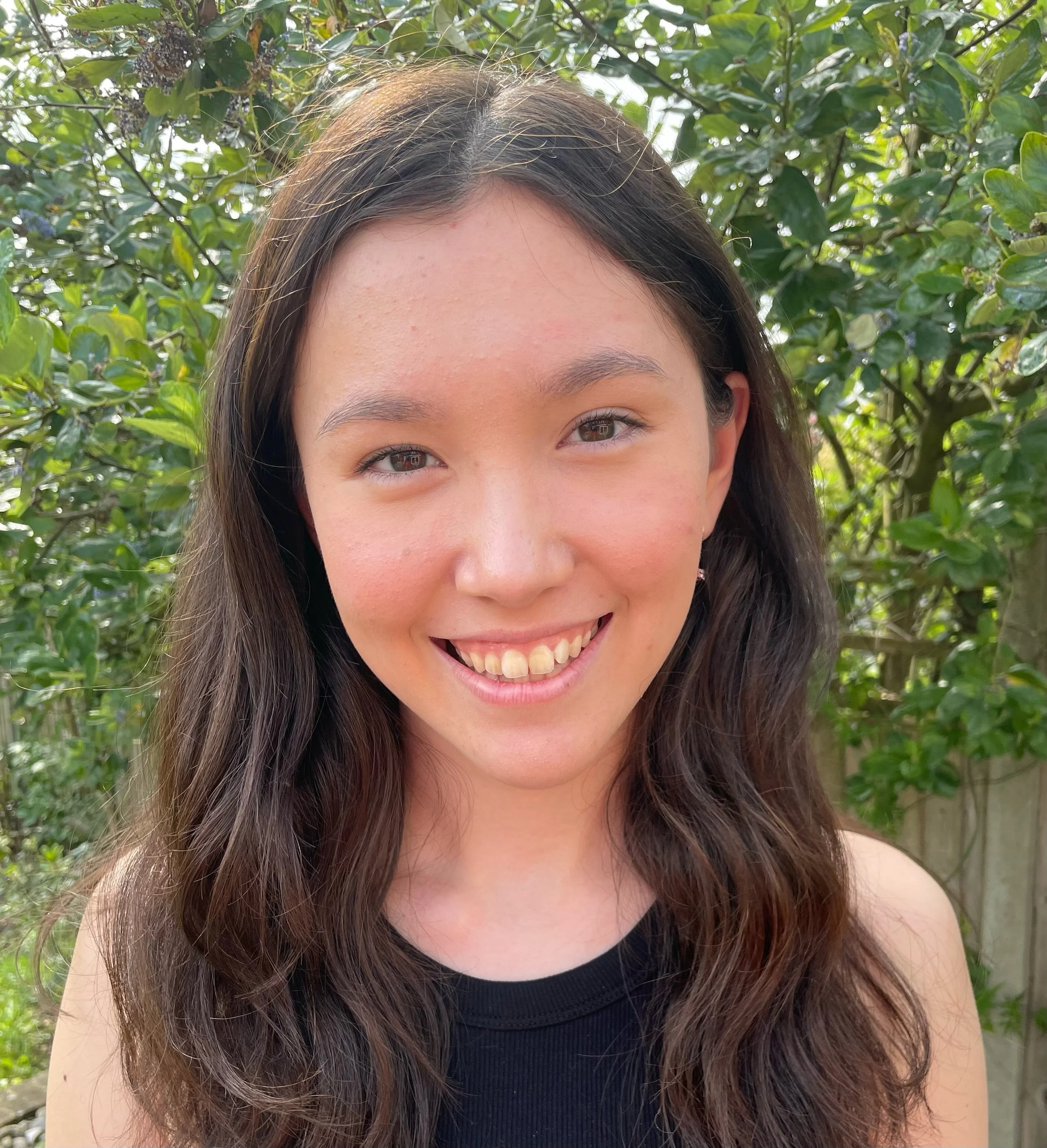 A young woman with long brown wavy hair smiling outdoors in front of green bushes.