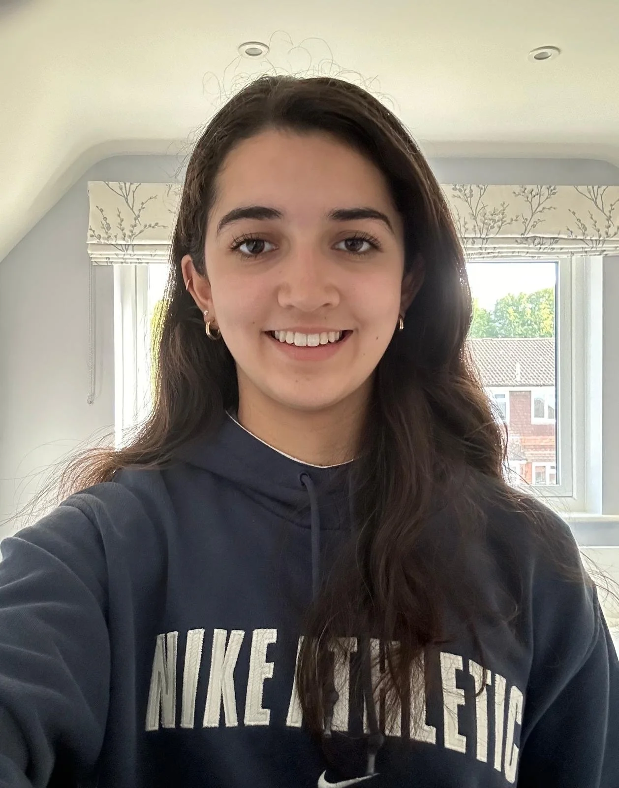 A young woman with long dark hair smiling and wearing a navy blue Nike hoodie, standing indoors with a window in the background showing a house and trees.