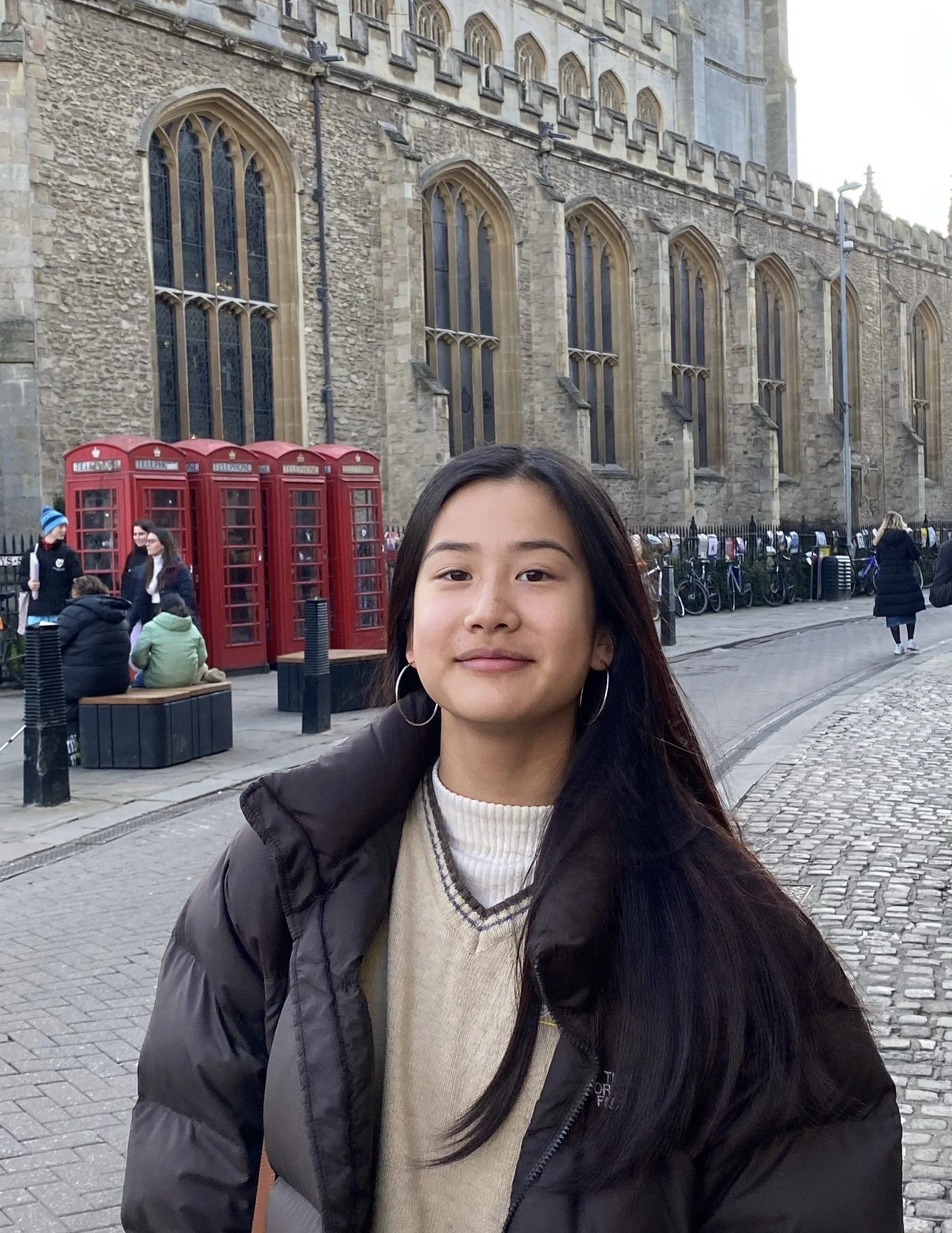 A young woman with long black hair, wearing hoop earrings, a beige sweater, and a black puffer jacket, stands on a cobblestone street in front of a stone church with tall pointed windows. Red telephone boxes and people sitting on benches are visible in the background.