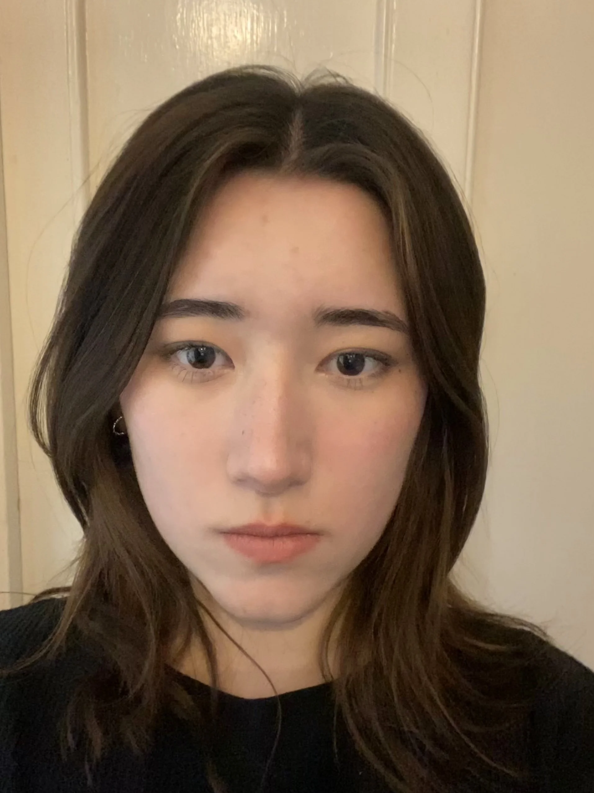 Close-up of a young woman with shoulder-length dark hair, wearing a black top, standing in front of a beige door.