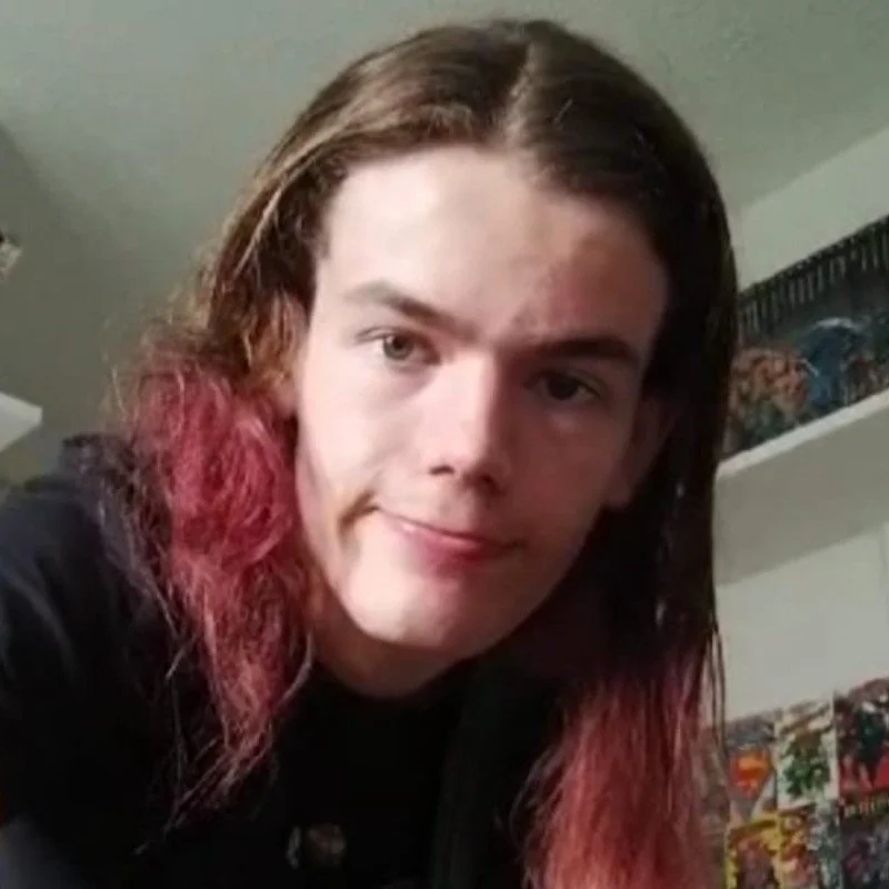 Young person with long hair that is brown at the top and red at the tips, smiling at the camera indoors with shelves of comic books or graphic novels in the background.