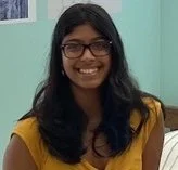 A woman smiling, wearing glasses and a yellow top, with dark hair, standing indoors against a light teal wall.