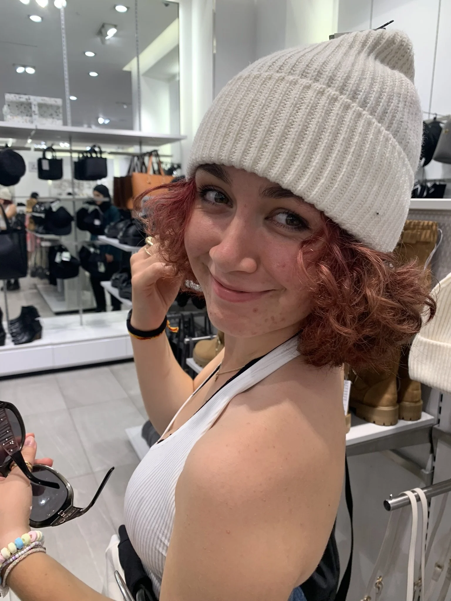 Young woman with curly red hair wearing a beige knit beanie and a sleeveless white top shopping in a store, holding sunglasses, with handbags on shelves in the background.