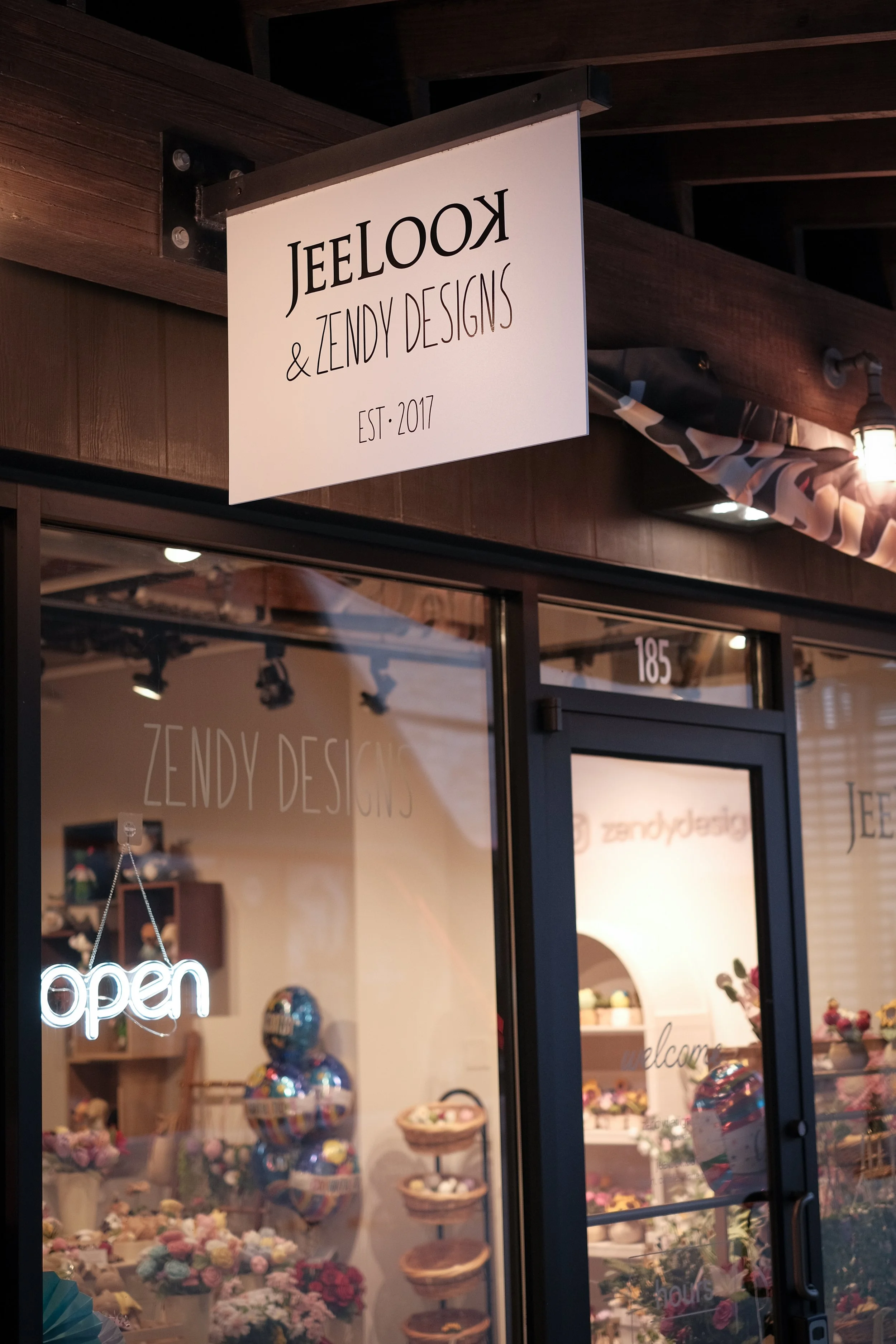 Storefront of JeelooK & Zendy Designs with a sign indicating it's established in 2017, a glass door with an 'open' sign, displaying flowers and balloons inside.