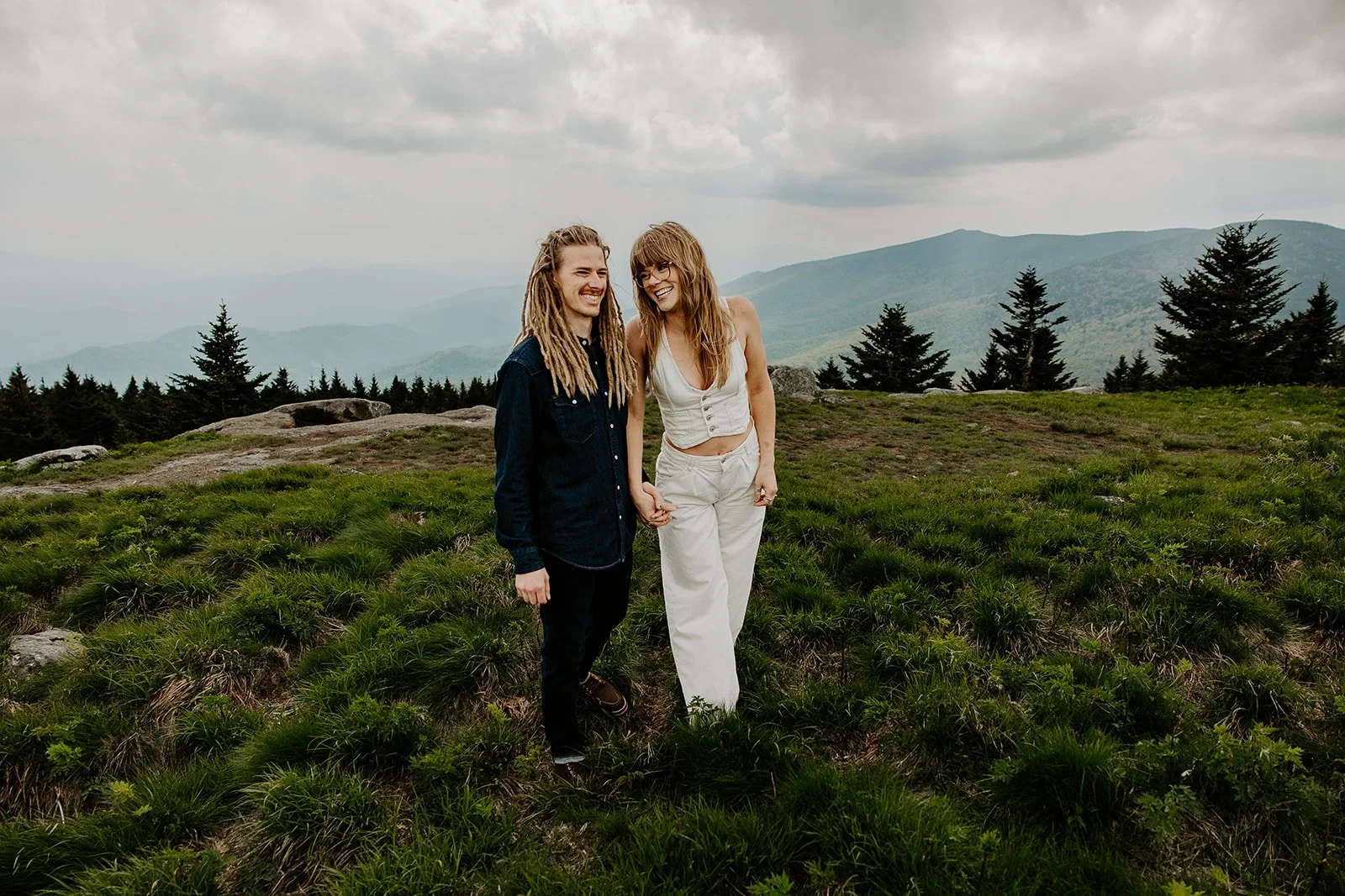 Couple in casual white wedding attire walking hand-in-hand on a lush West Virginia mountain summit with cloudy Appalachian mountain views
