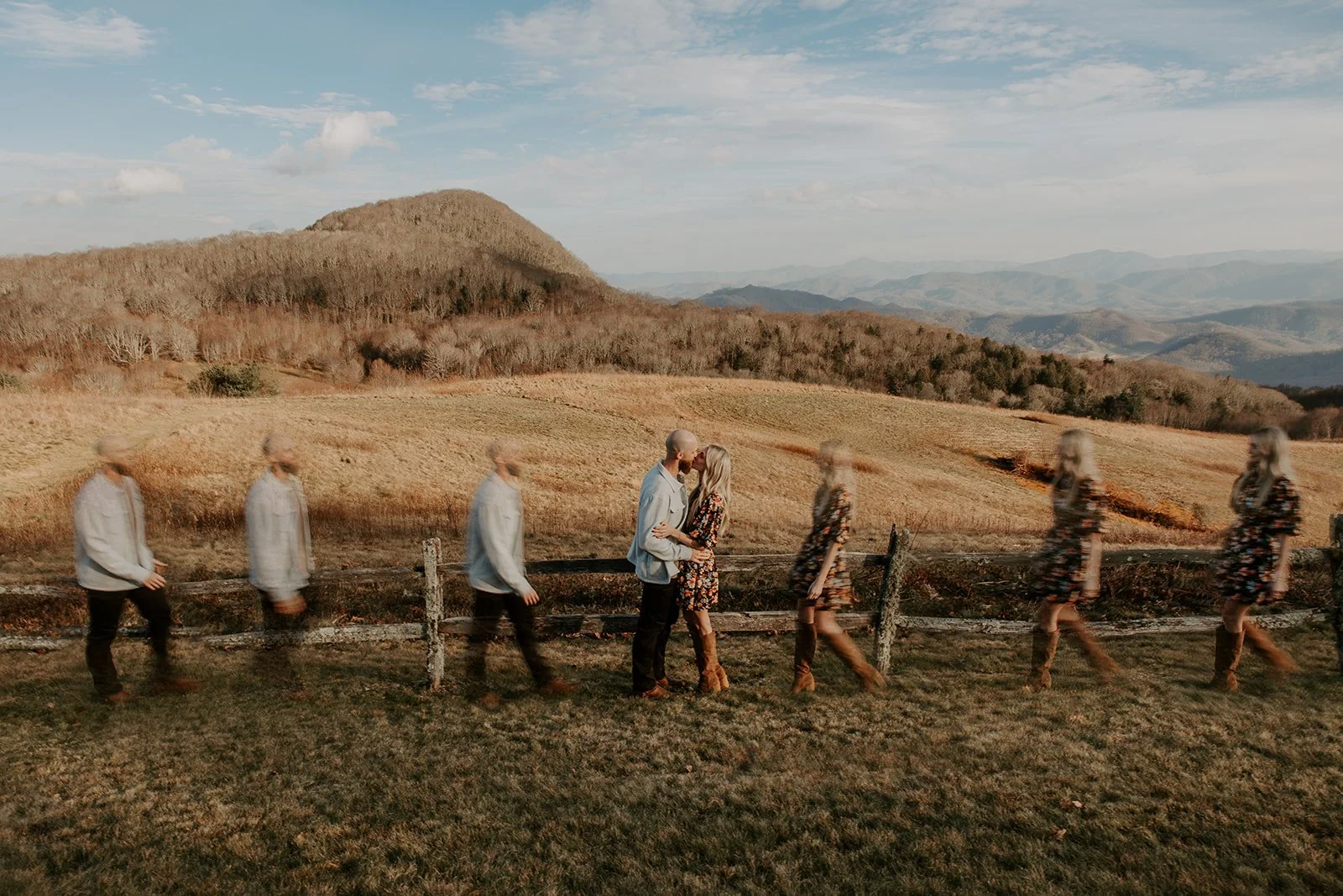 Couple kissing at center while motion-blurred figures walk past along a split rail fence at Purchase Knob with sweeping Smoky Mountain views behind them