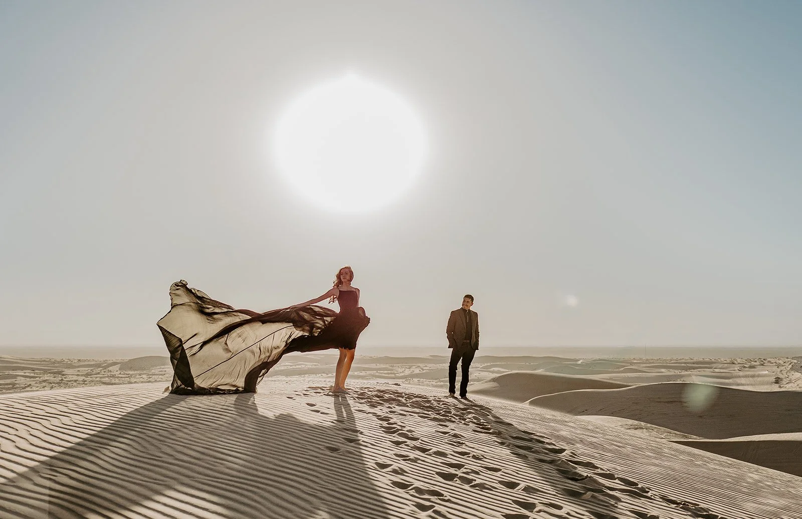 Bride standing on a sand dune with a flowing dress blowing dramatically in the wind while her partner looks on