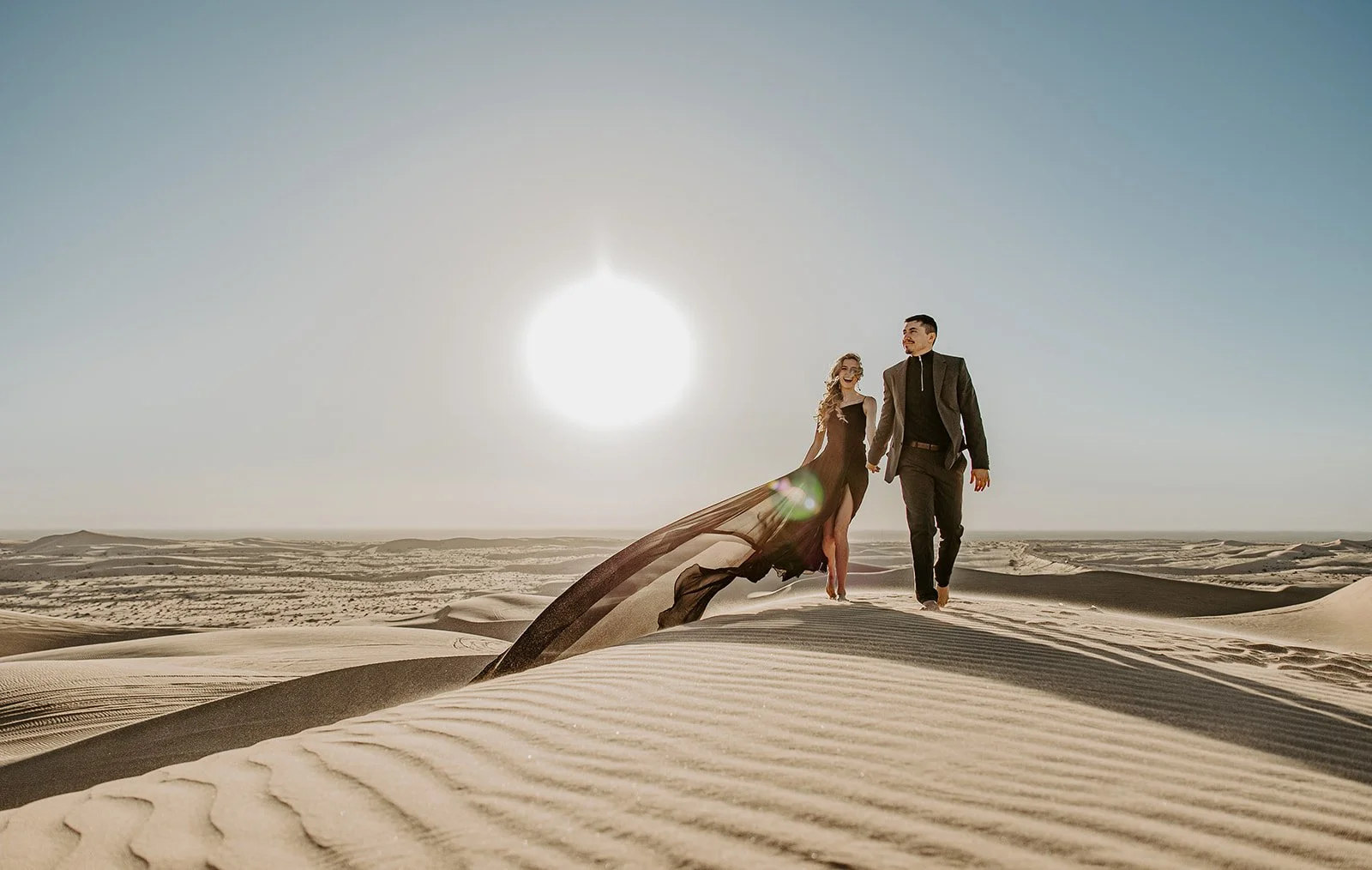 Couple walking across a sunlit sand dune with a flowing dress trailing behind them