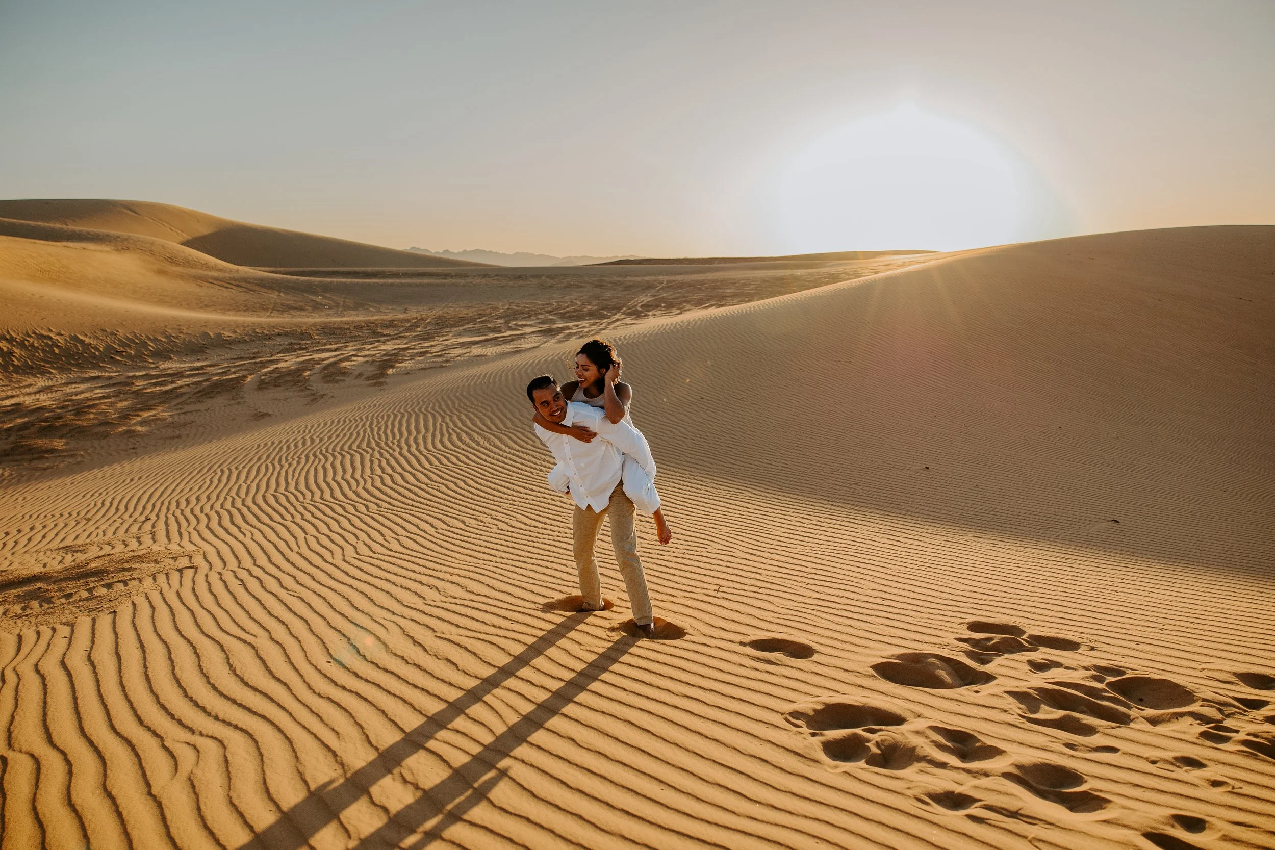 Couple laughing during a playful piggyback moment on rippled sand dunes at sunrise