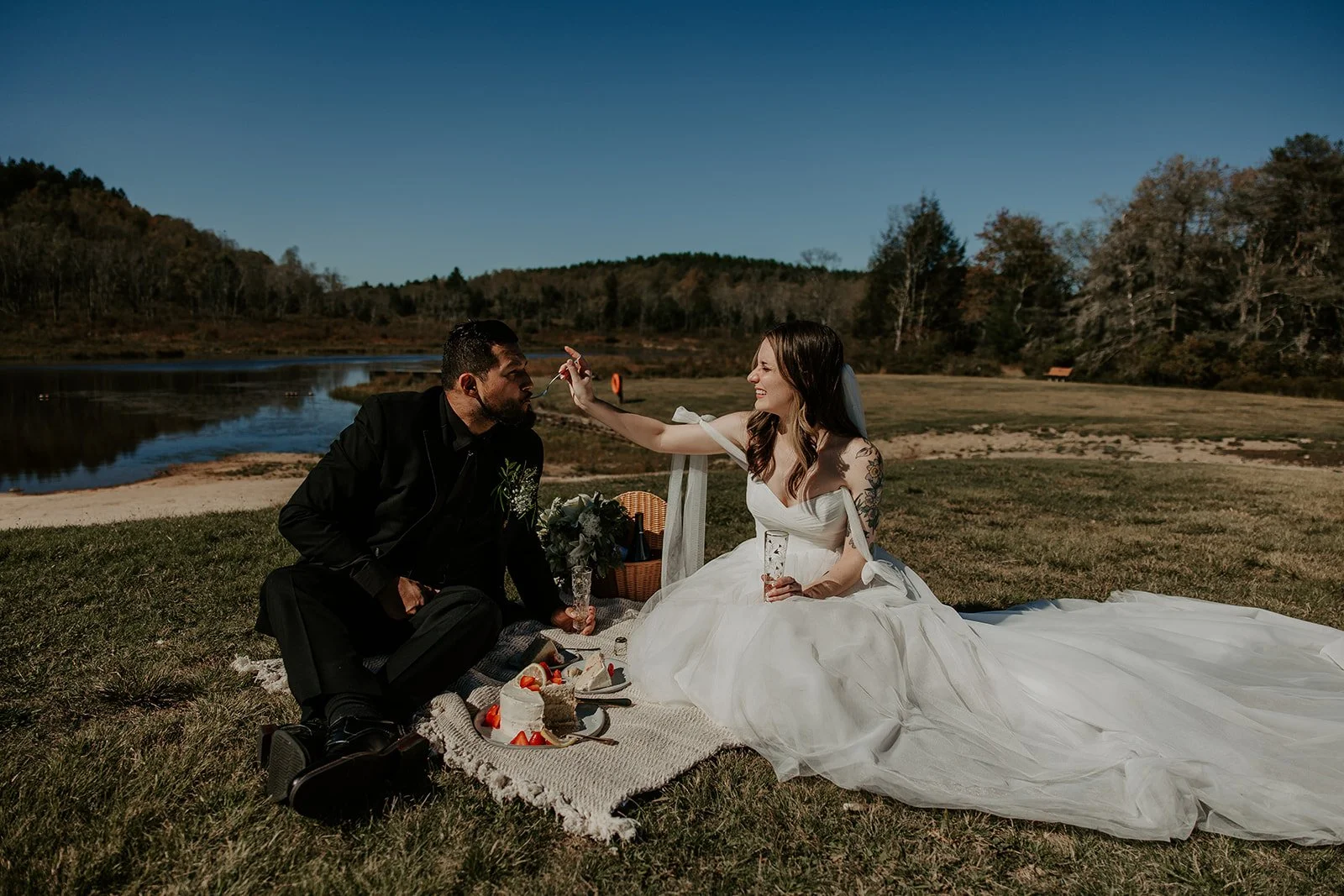 Bride and groom in wedding attire celebrating their West Virginia elopement with a lakeside picnic, wedding cake, strawberries, and champagne