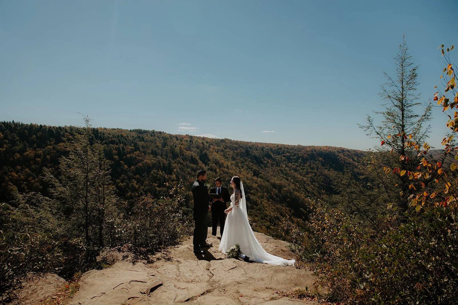 Couple exchanging vows with an officiant on the rocky outcrop at Pendleton Point Overlook in Blackwater Falls State Park, West Virginia, with a forested gorge and early fall foliage stretching behind them