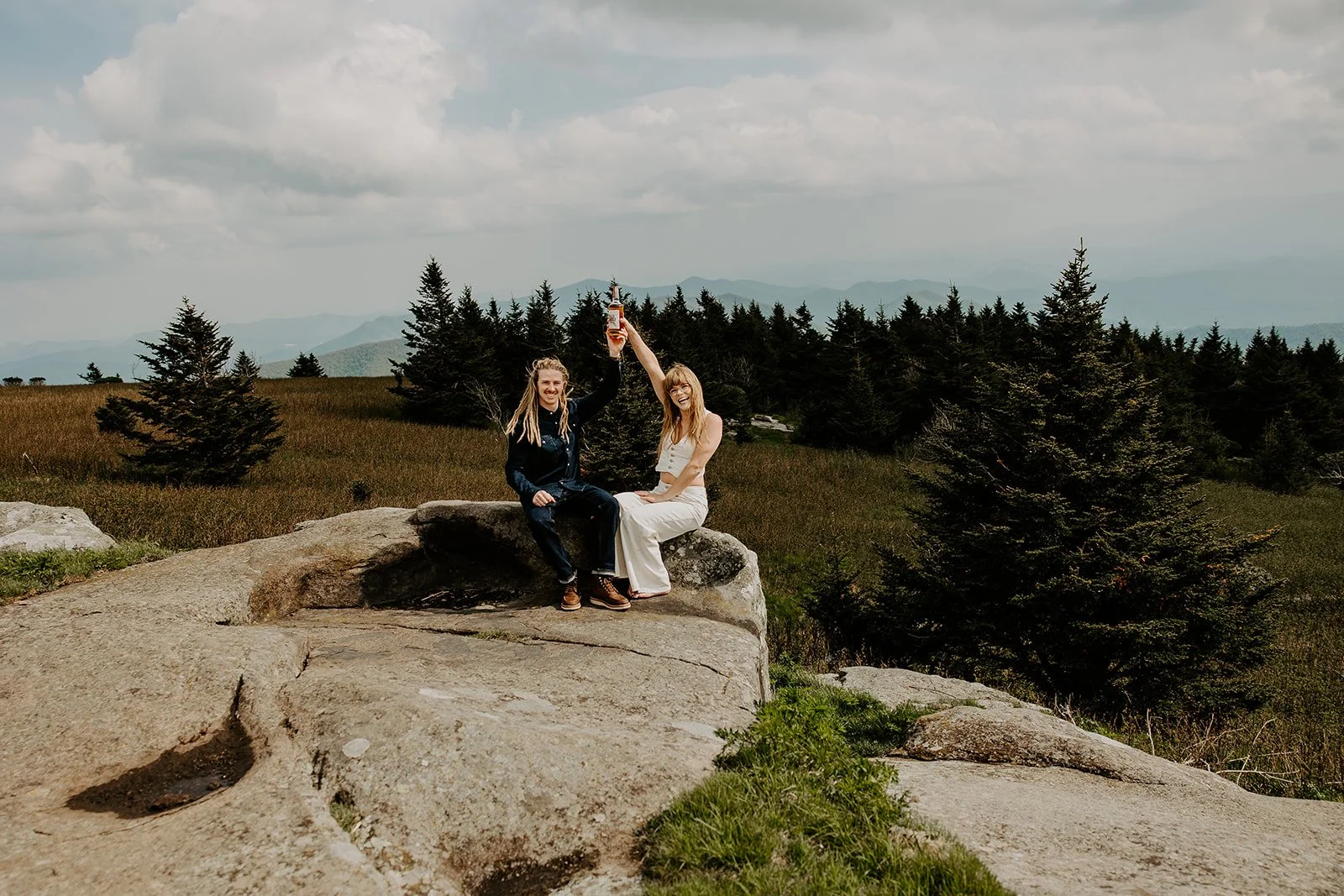 Couple sitting on a large granite boulder on a high-elevation Appalachian mountain bald, raising a bottle in celebration, with spruce trees and open mountain meadow behind them