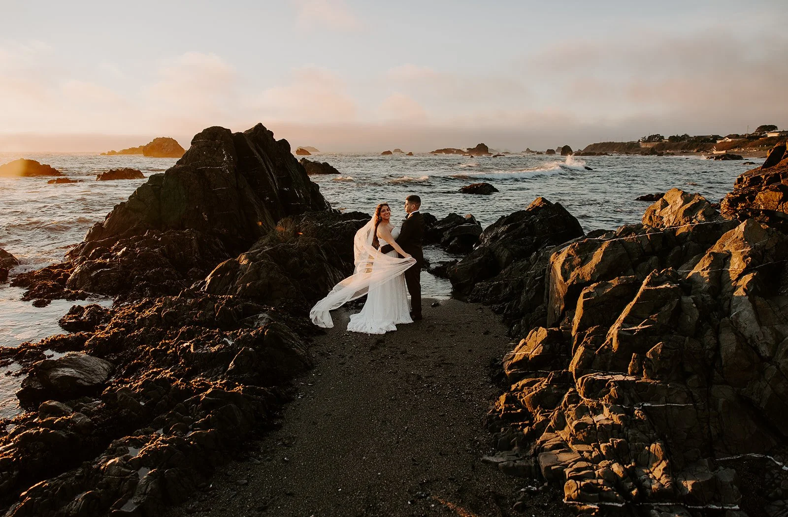 Elopement couple on the rocky shoreline at Battery Point Beach, Crescent City, California at golden sunset, veil flowing in coastal wind near the northern redwoods