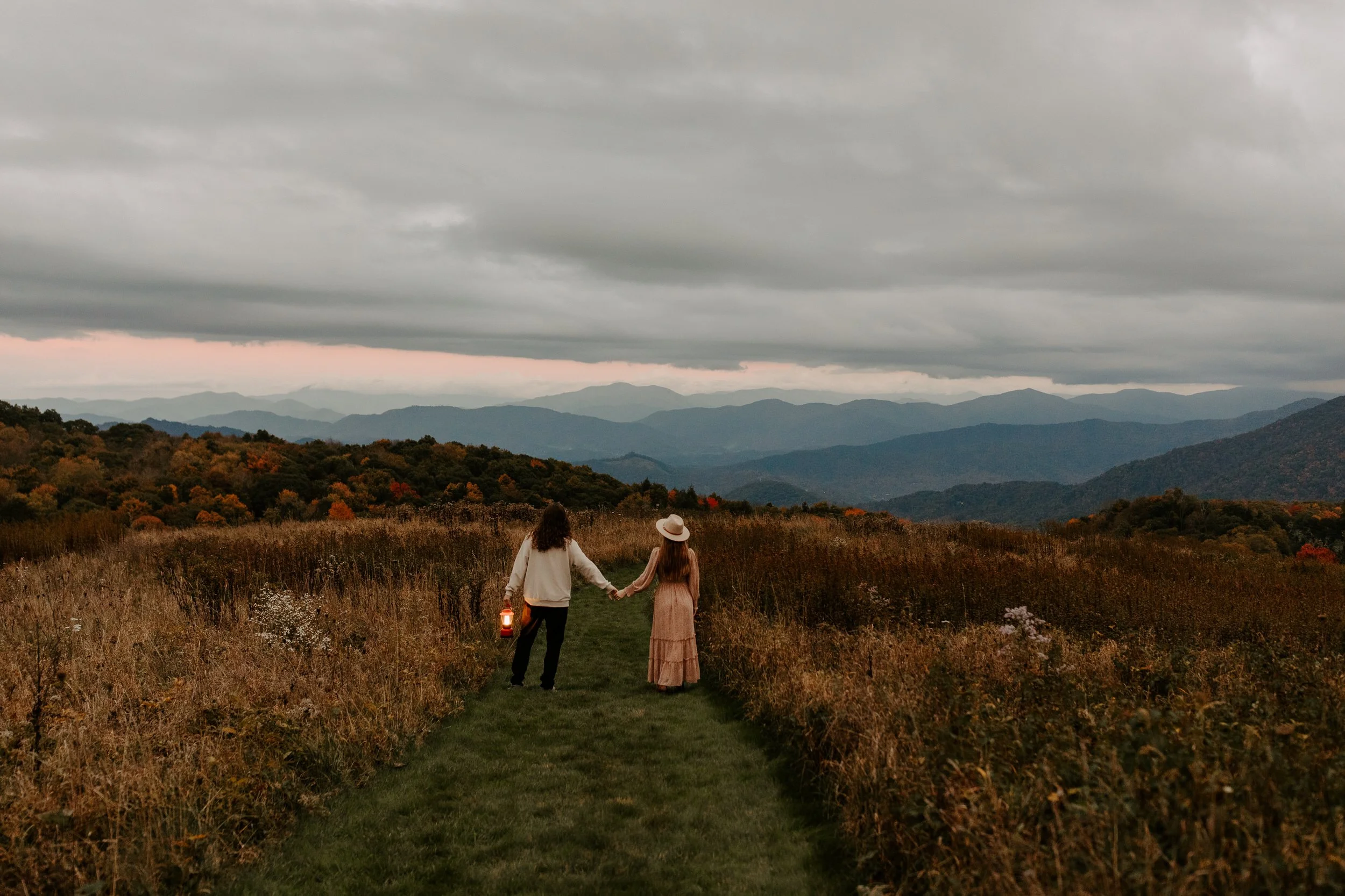 Couple walking hand in hand down a grass path through golden fall meadow with Smoky Mountain ridgelines in the distance, one partner holding a red lantern