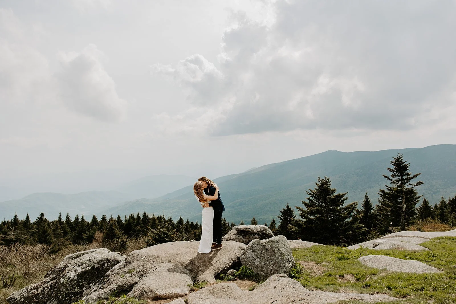 Couple embracing on a rock outcrop at a high-elevation Appalachian bald with lush green spruce trees and layered mountain ridges under cloudy skies
