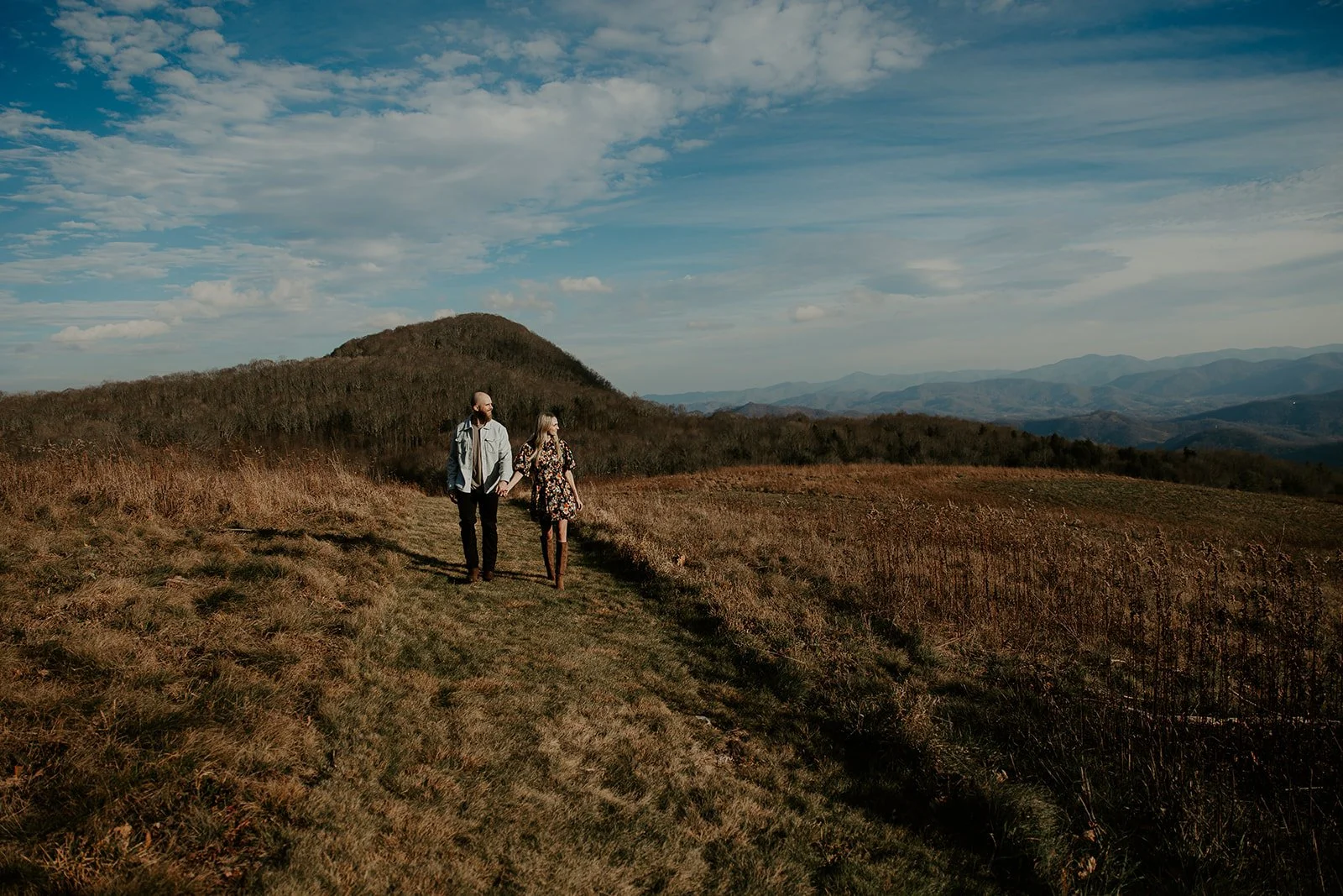 Couple walking hand in hand along a grassy ridge at Purchase Knob with a rounded mountain summit and Smoky Mountain layers extending to the horizon