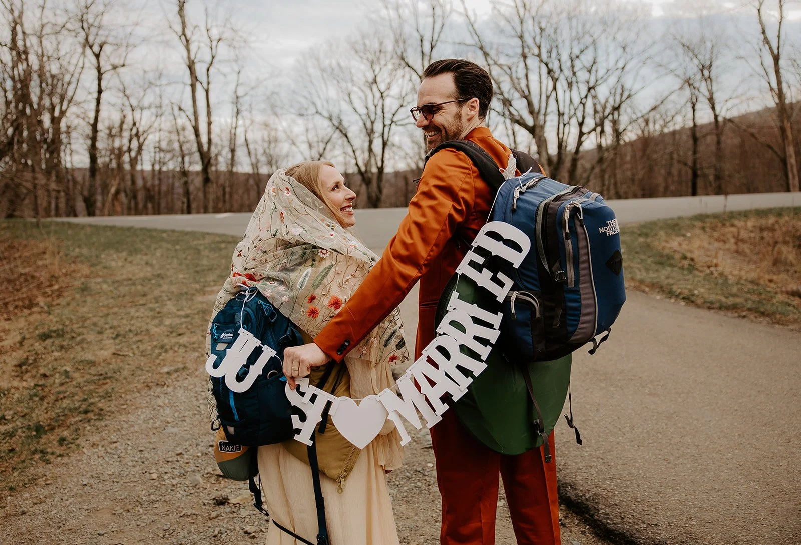 Newlyweds carrying hiking backpacks and holding a Just Married banner after eloping in West Virginia, wearing bold colorful wedding attire on a mountain road
