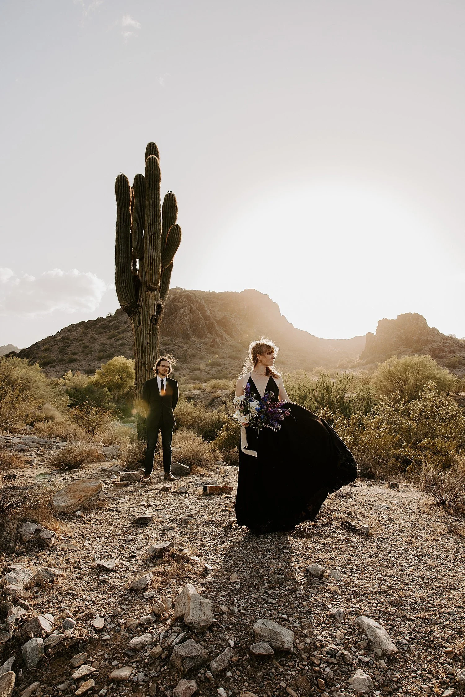 Groom standing off in the distance admiring his bride standing in front of him playing with the skirt of her black wedding dress and holding her blue, purple, pink, and white bouquet.