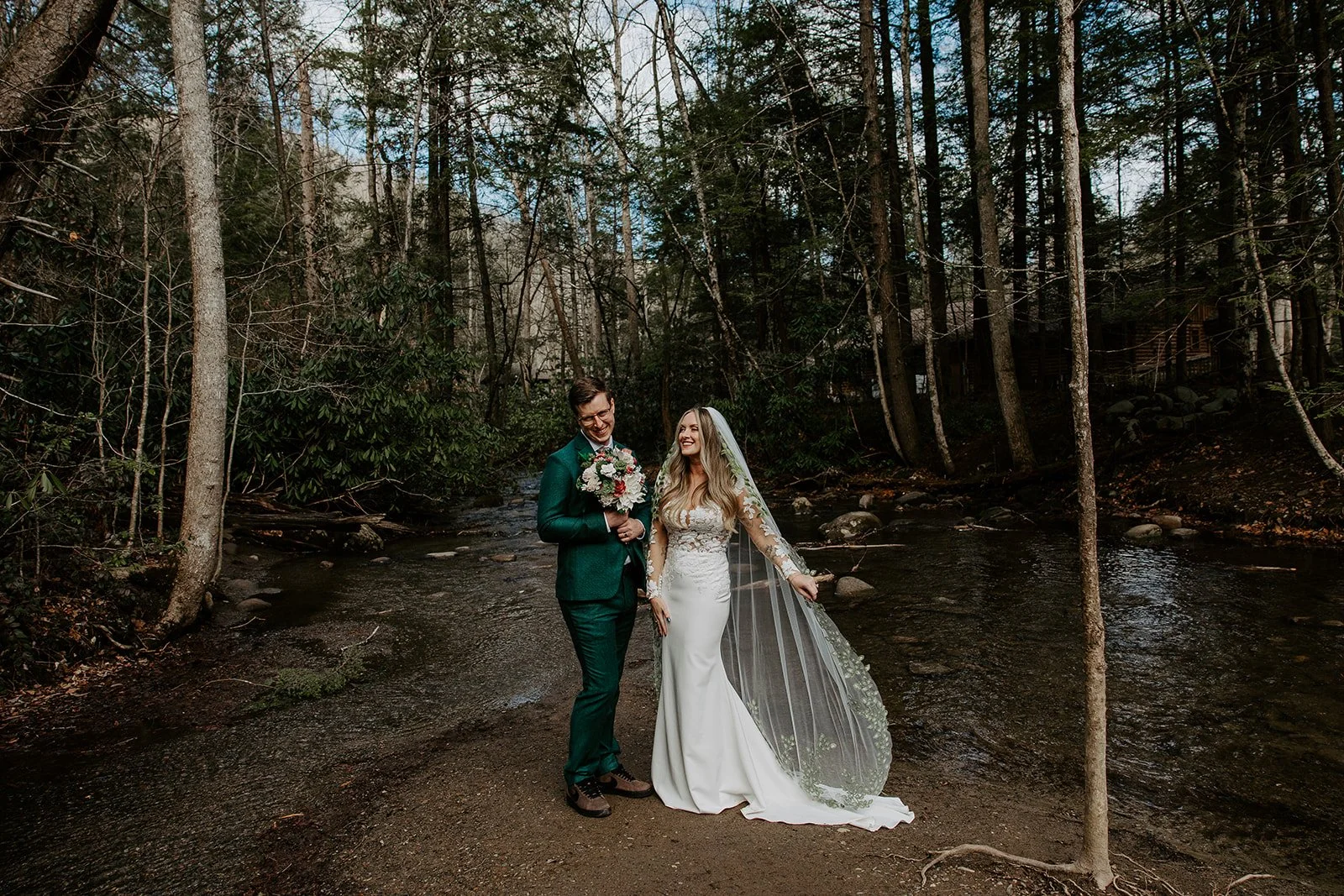 Bride in lace veil gown and groom in emerald suit walking along a rocky creek in winter forest in Great Smoky Mountains National Park