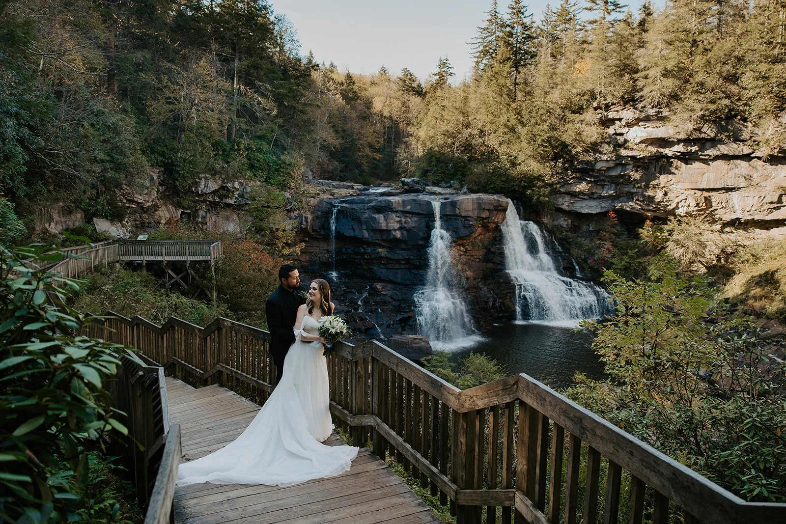 Couple standing on the wooden overlook deck at Blackwater Falls State Park during their West Virginia elopement, bride in an off-the-shoulder gown with a long train, waterfall and fall foliage in the background