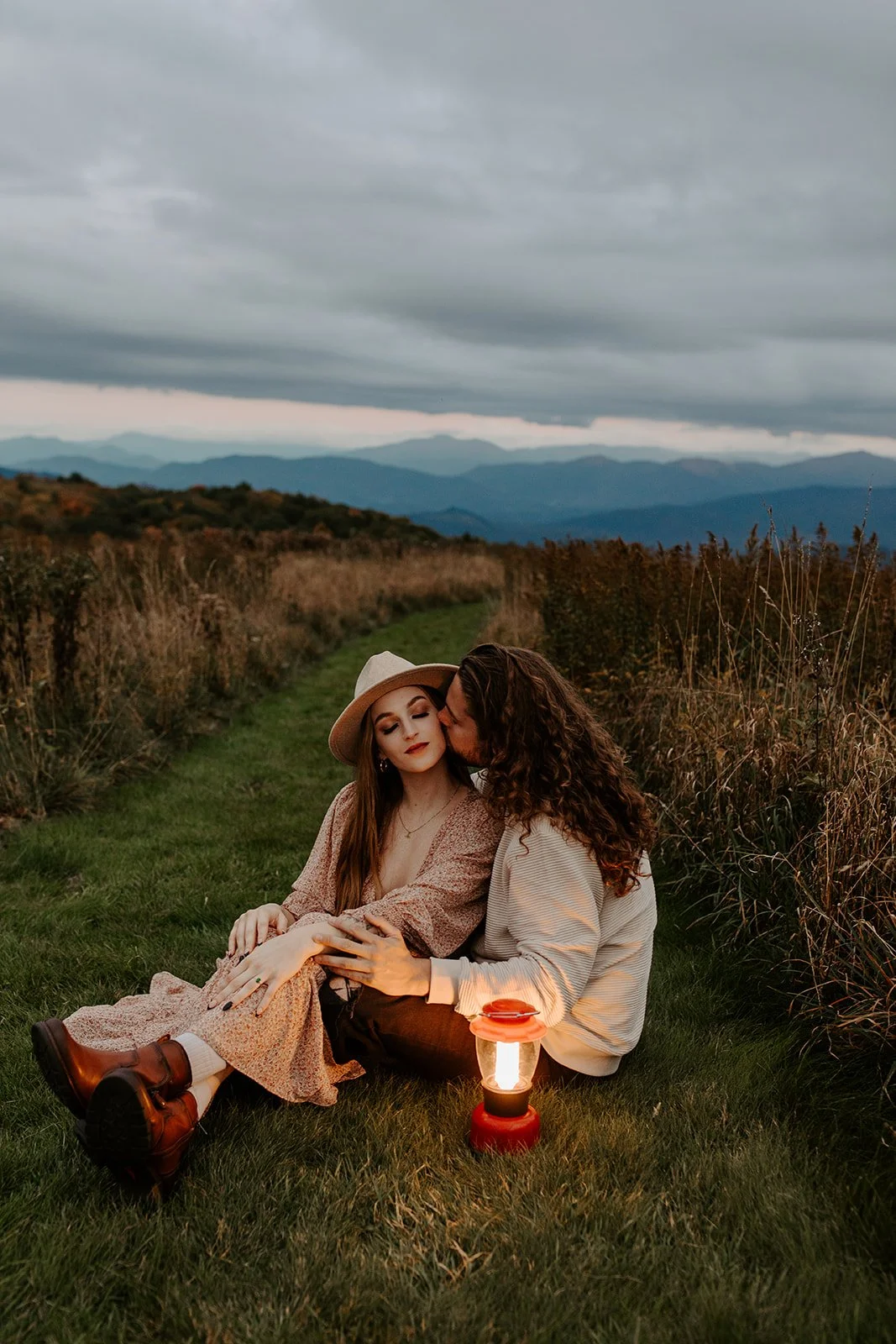 Couple sitting together on a grass path at blue hour with a glowing red lantern, Smoky Mountain ridgelines fading into the dusky sky behind them