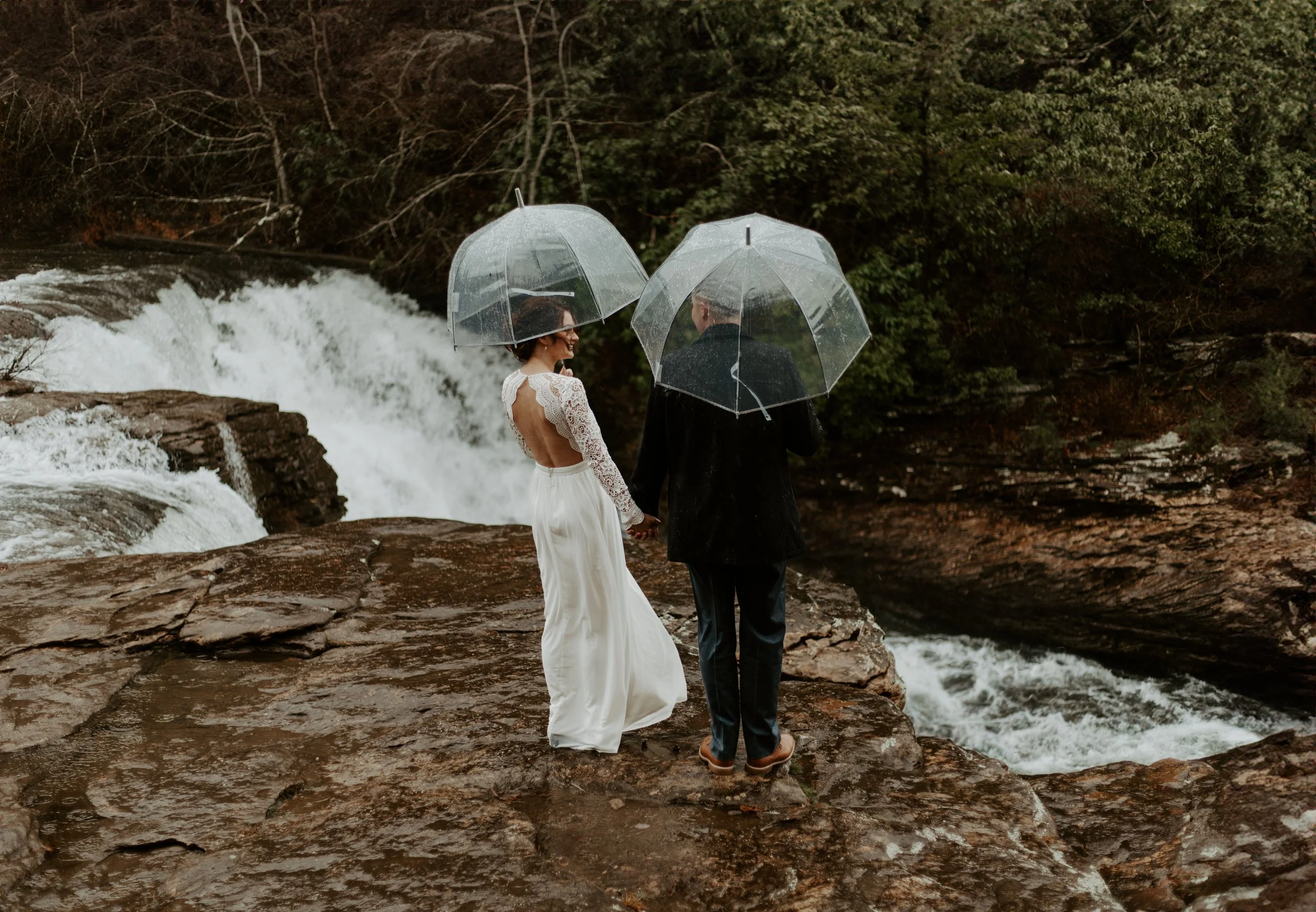 Couple holding hands under clear umbrellas beside DeSoto Falls during a rainy waterfall elopement