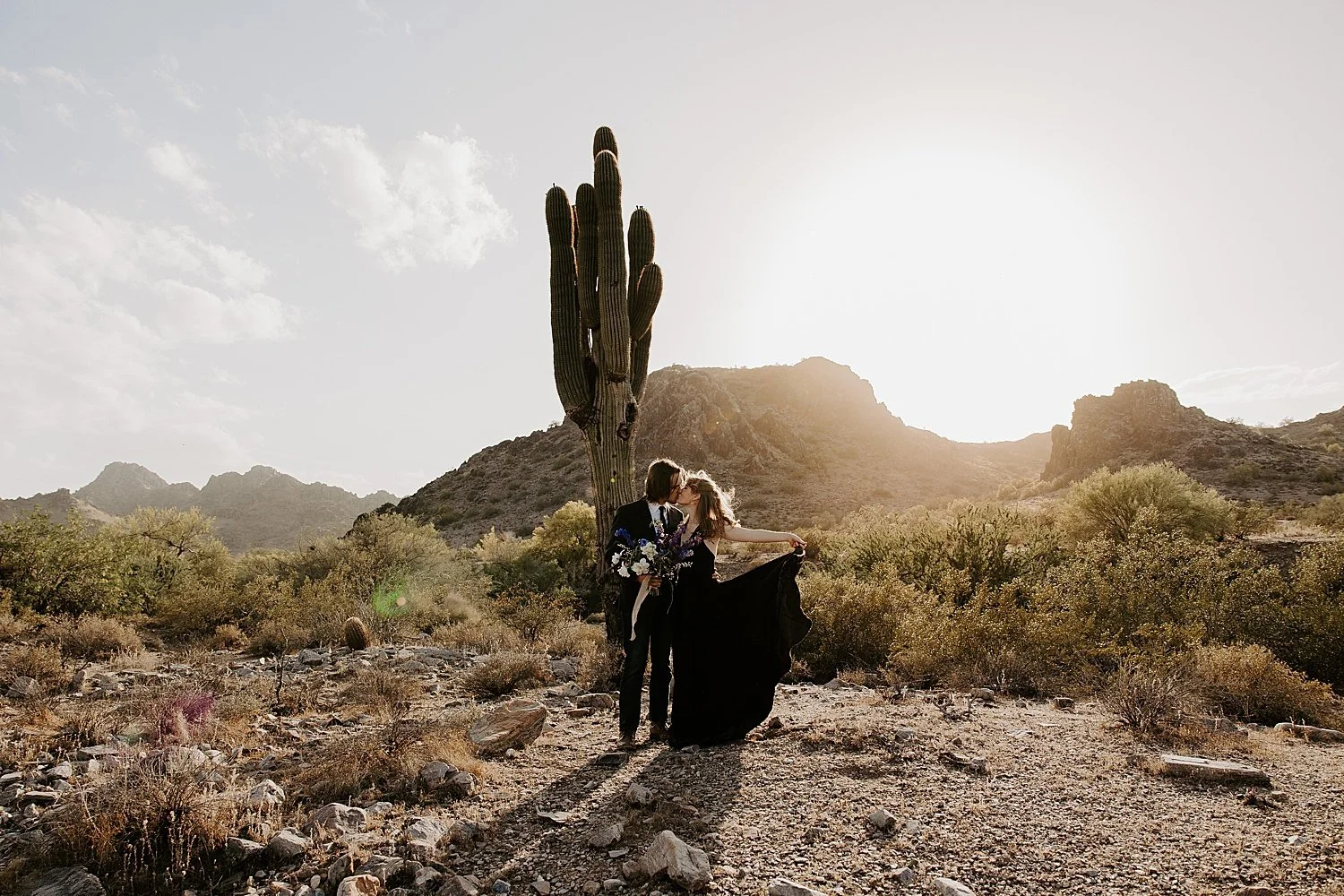 Bride and groom in all black wedding attire with the groom holding the blue, purple, pink, and white bridal bouquet as the bride swings her dress out beside her in the desert.