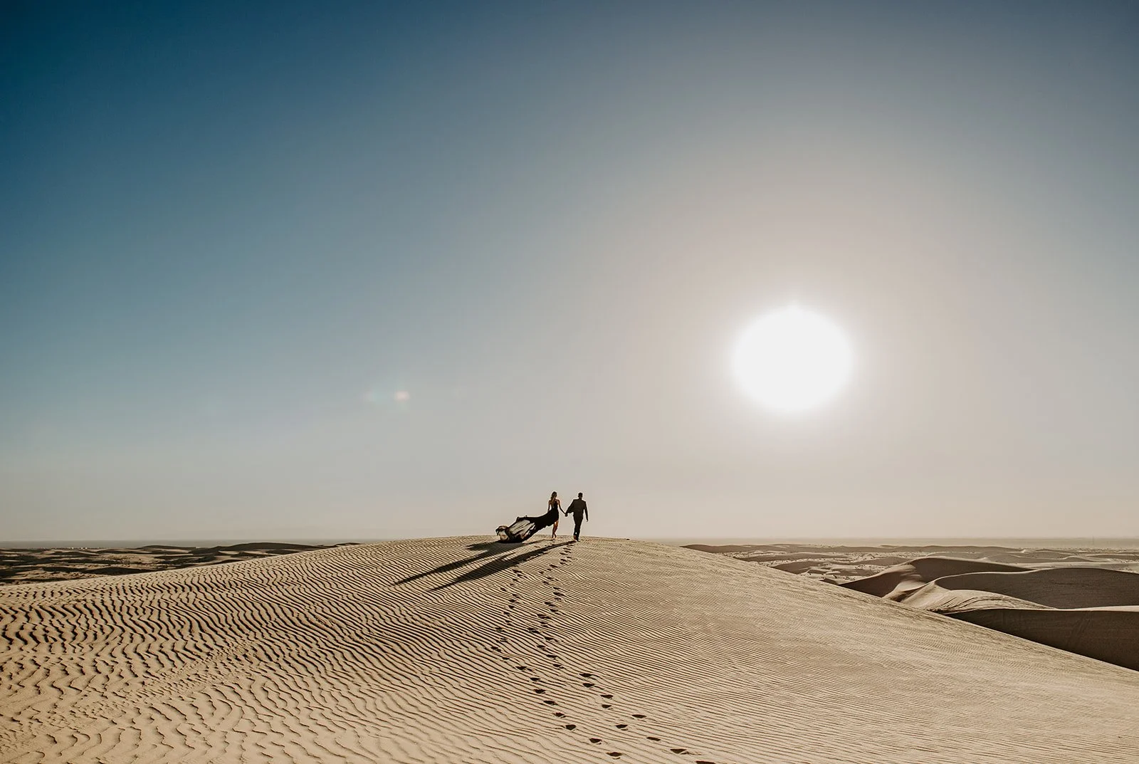 Couple walking along the top of a sand dune ridge with the sun overhead and desert dunes stretching into the distance