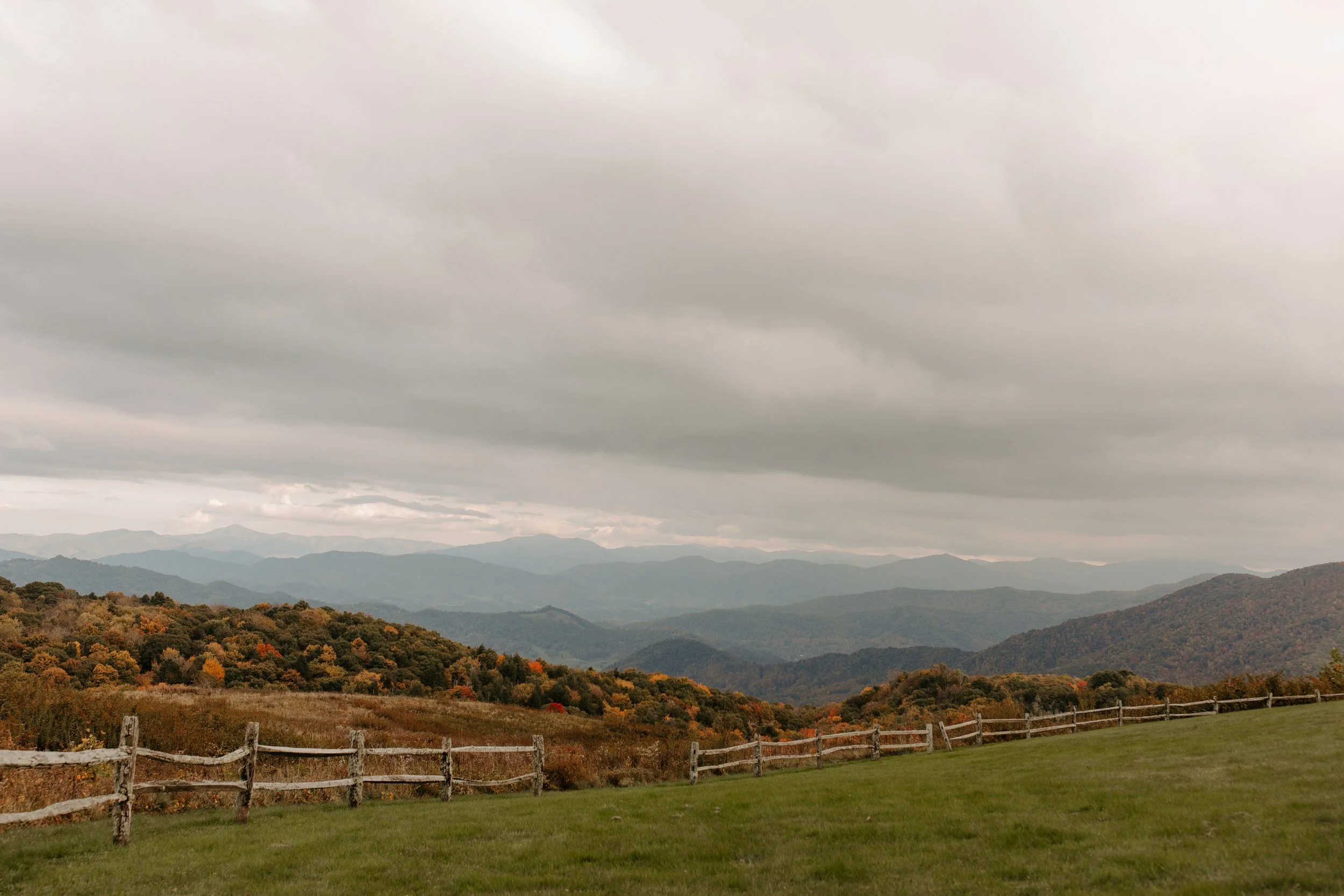 Panoramic autumn view from Purchase Knob in the Smoky Mountains with split rail fence, golden meadow, and layered blue ridgelines under a cloudy sky
