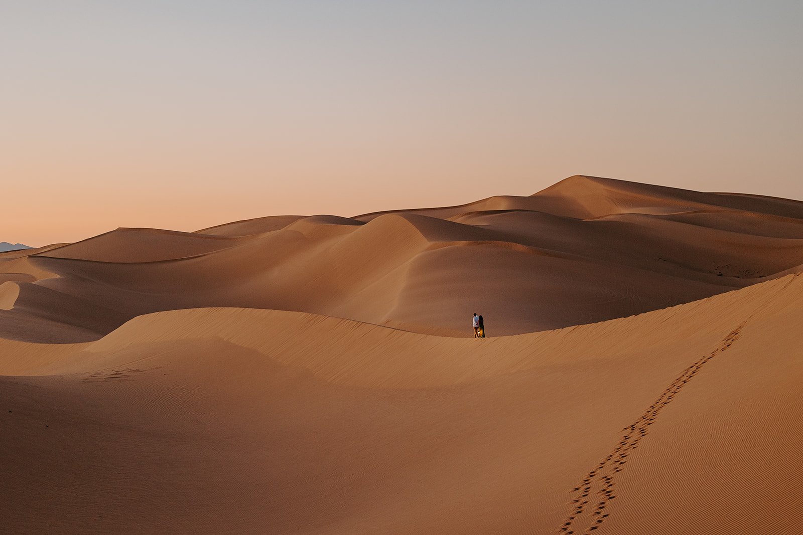 Couple standing together in the distance on sweeping sand dunes at sunrise with footprints leading across the ridge