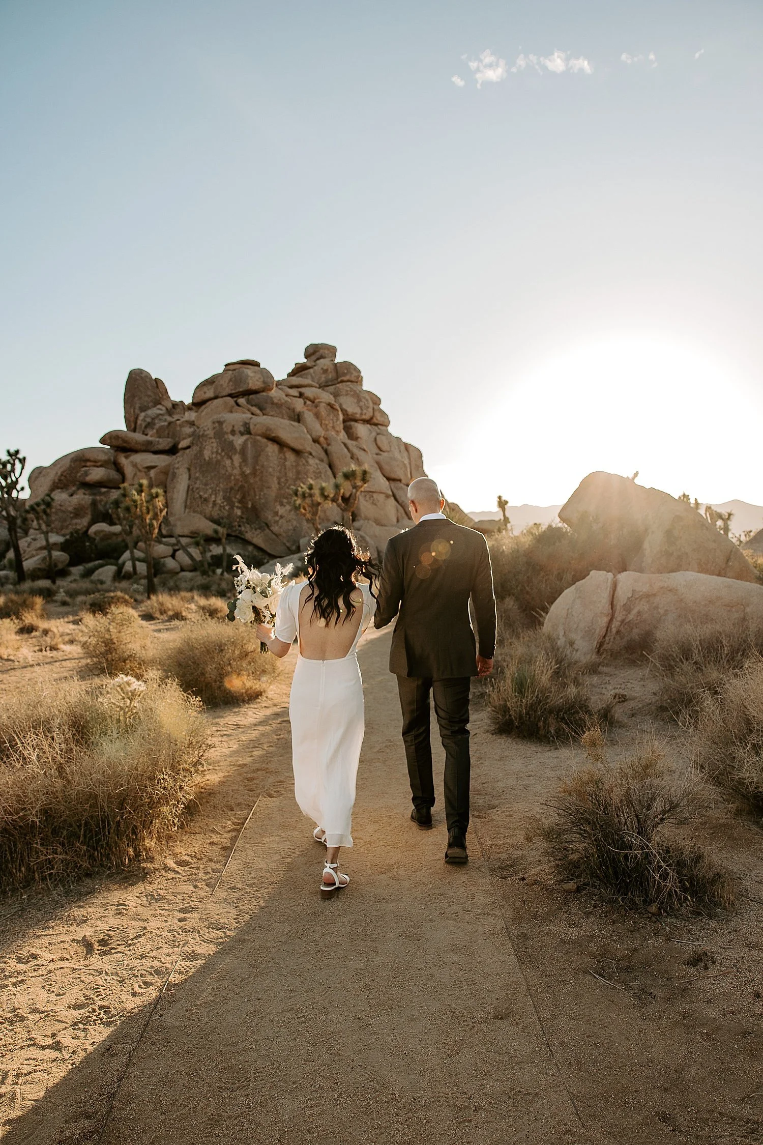 Bride and groom walking down a trail and off into the sun as it sinks lower in the sky at Cap Rock in Joshua Tree National Park
