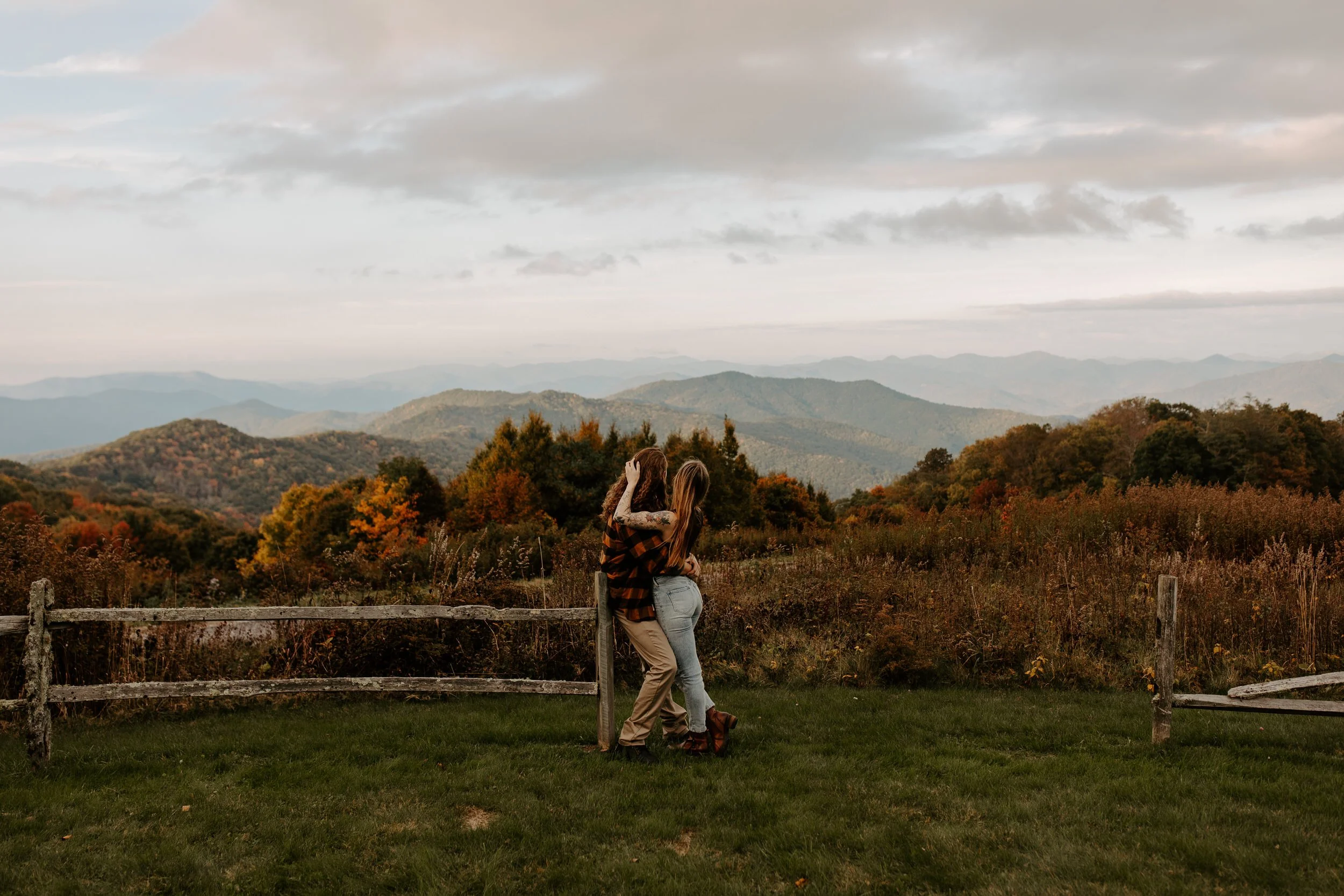 Couple leaning together against a weathered split rail fence overlooking early fall foliage and layered Smoky Mountain ridgelines at dusk