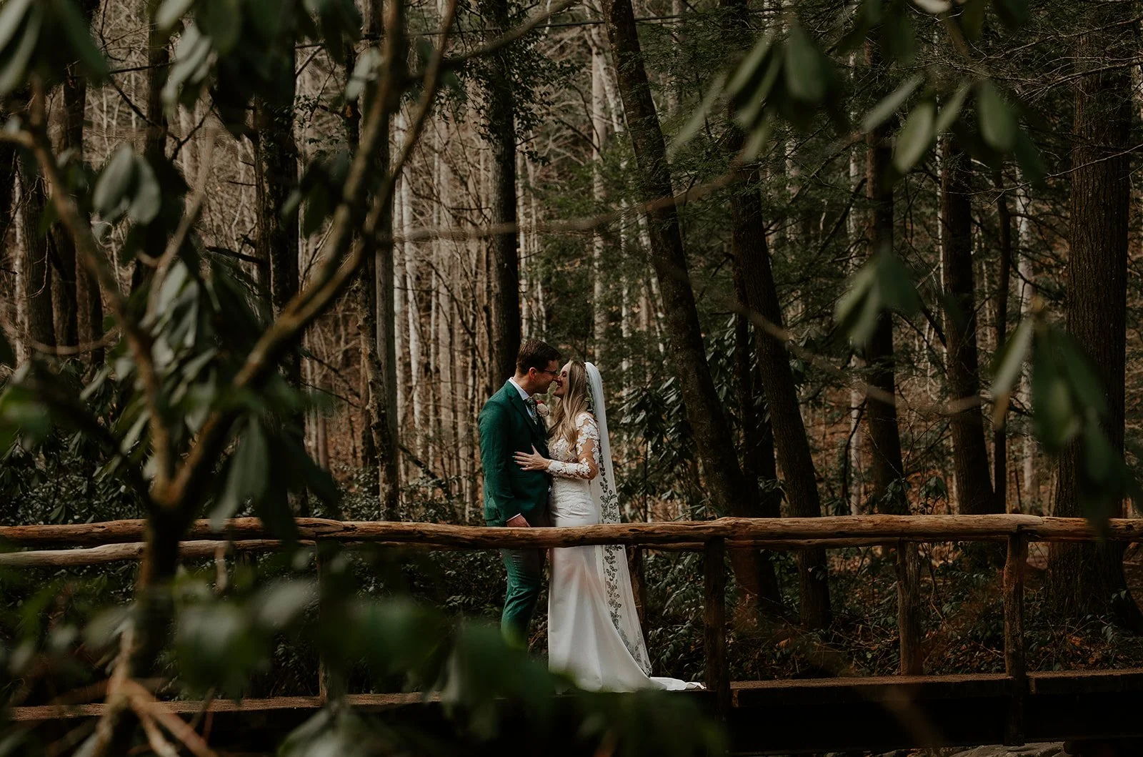 Bride in a white lace gown and groom in a green suit sharing a moment on a rustic wooden bridge surrounded by a dense West Virginia forest