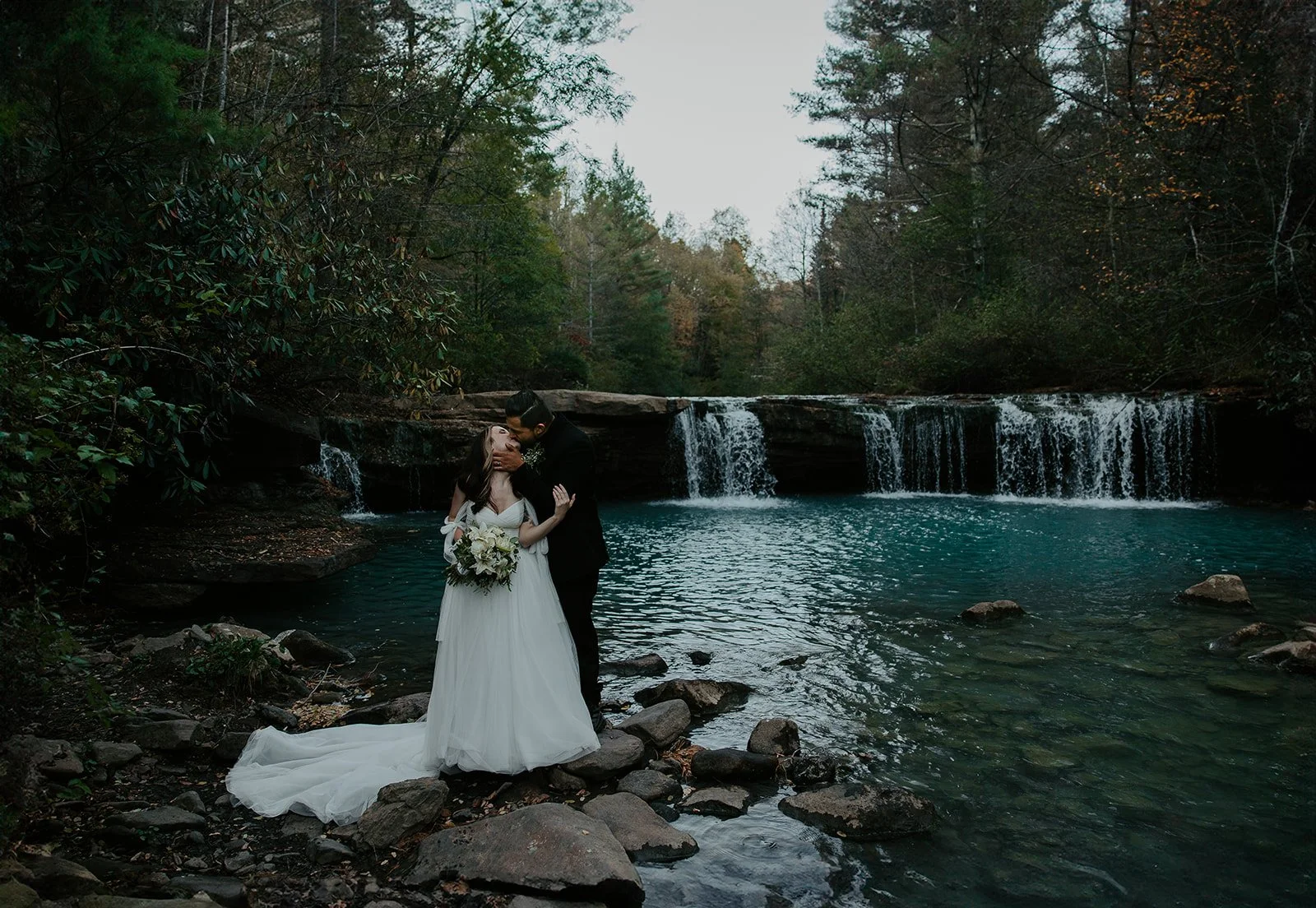 Couple kissing on the rocks beside the teal pool at Albert Falls in West Virginia, surrounded by dark forest, rhododendron, and late fall foliage
