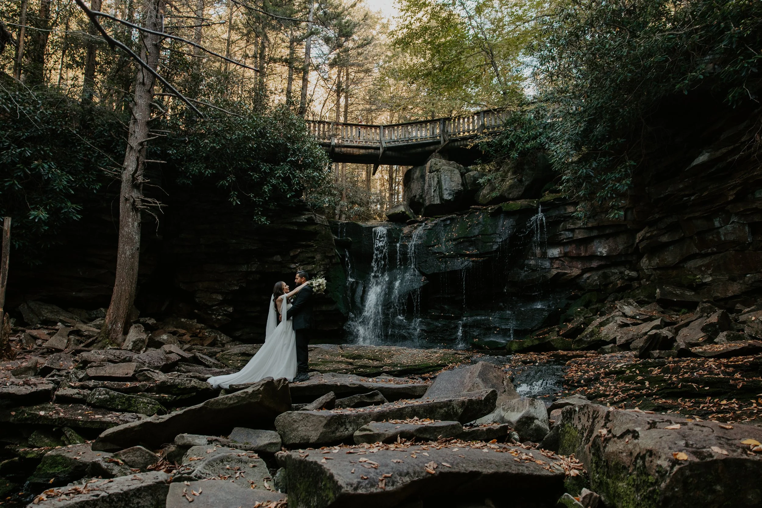 Couple embracing at the base of Elakala Falls in Blackwater Falls State Park with a wooden bridge overhead and fall leaves scattered across the rocks