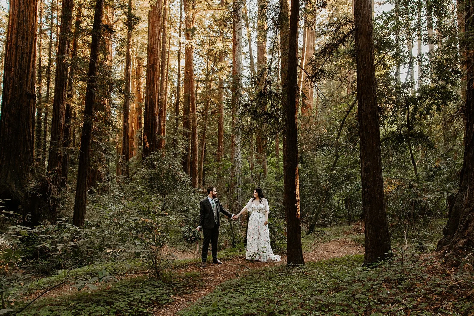 Couple holding hands facing each other on a warm sunlit trail in the Redwood Grove, Henry Cowell Redwoods State Park, bride in a floral gown, California elopement