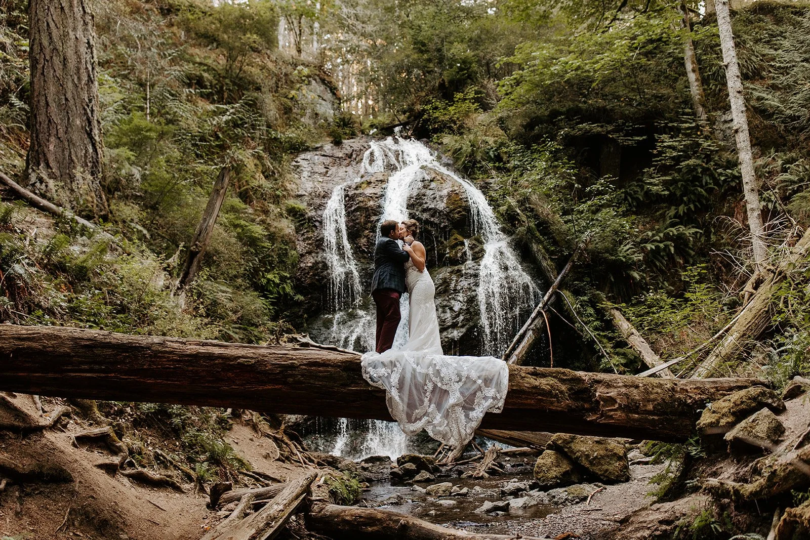 Bride and groom kissing on fallen log beneath Cascade Falls in Moran State Park.